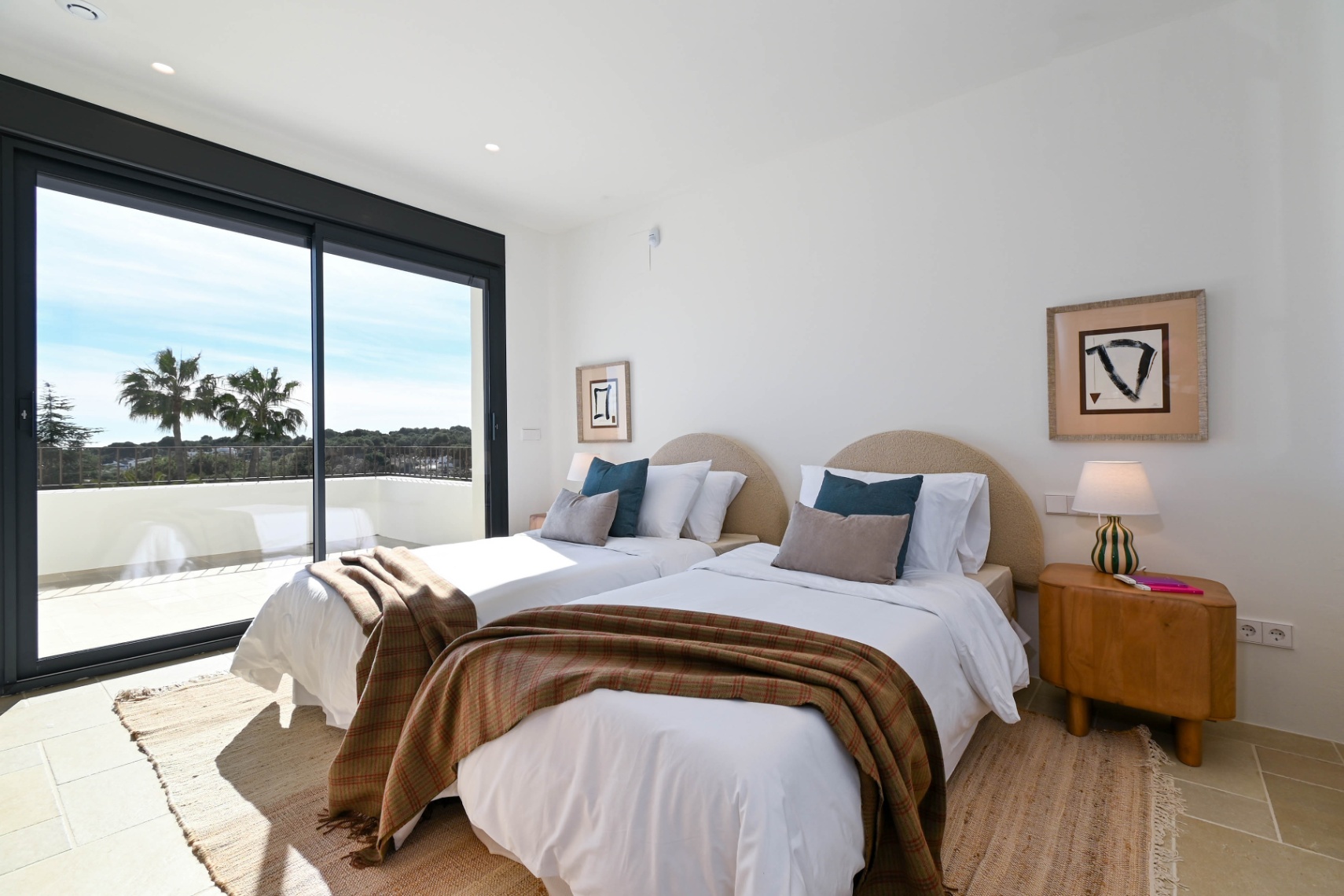 Helles Schlafzimmer mit zwei Einzelbetten, geschwungenen Kopfteilen und direktem Zugang zur Terrasse mit Blick auf Palmen und die umliegende Landschaft.