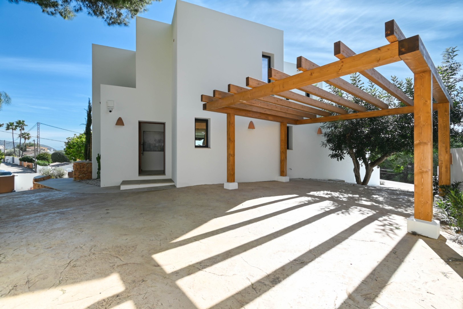Minimalistische weiße Fassade mit einer großen Pergola aus Naturholz über einem gepflasterten Parkplatz. Mit keramischen Wandleuchten und modernem Eingang.