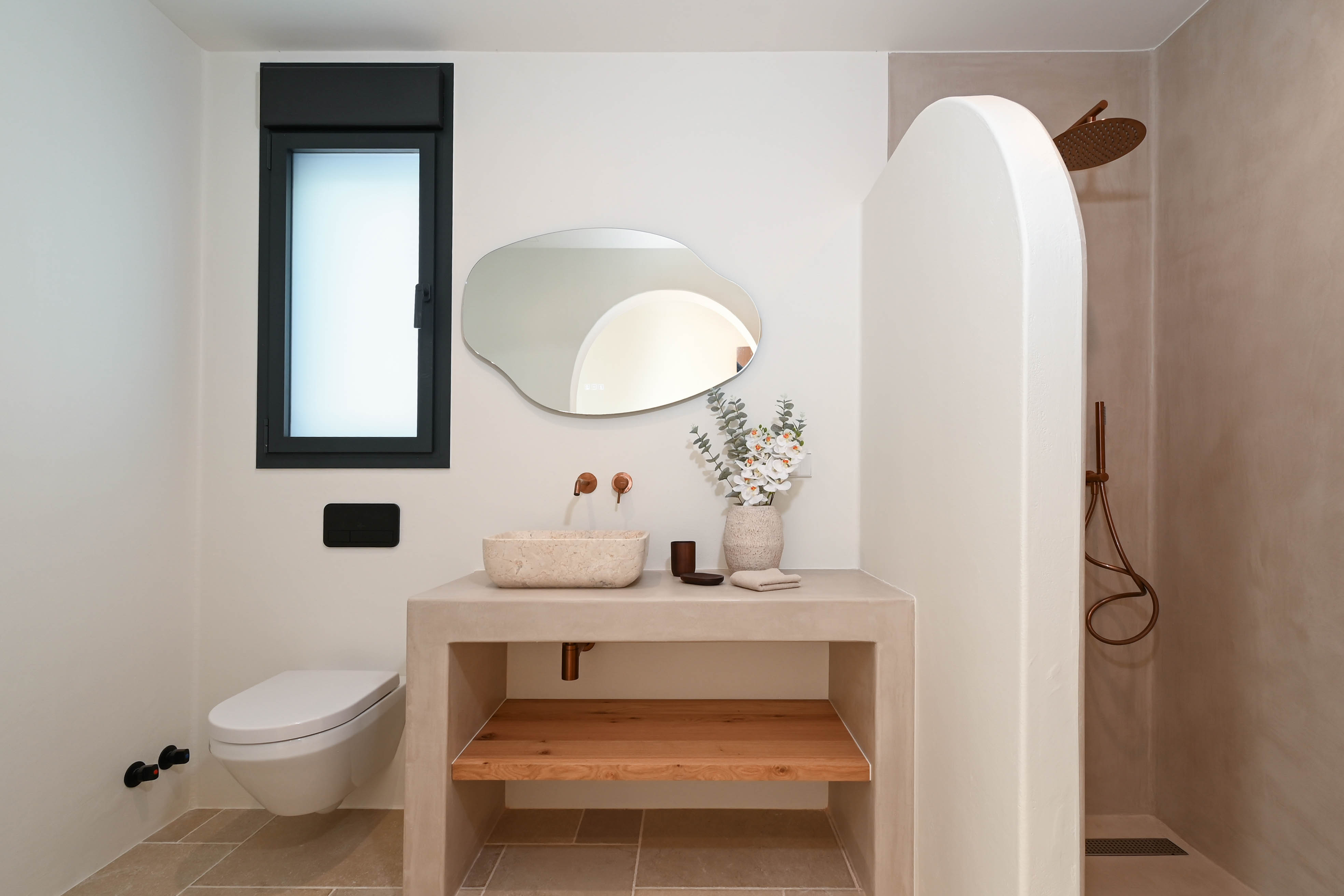 Modernes Badezimmer mit Mikrozement-Waschtisch, Natursteinbecken und Kupferarmaturen. Es bietet einen organisch geformten Spiegel, ein Wand-WC und eine Dusche mit geschwungener Trennwand.