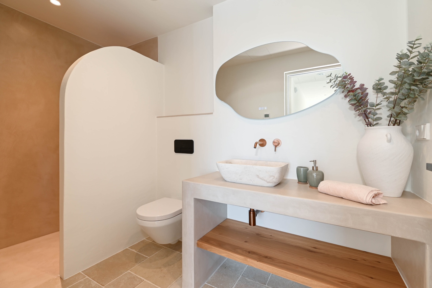Modernes Badezimmer mit Steinwaschbecken auf Mikrozement-Waschtisch, wandmontierten Kupferarmaturen, organisch geformtem Spiegel und einer geschwungenen Duschtrennwand.