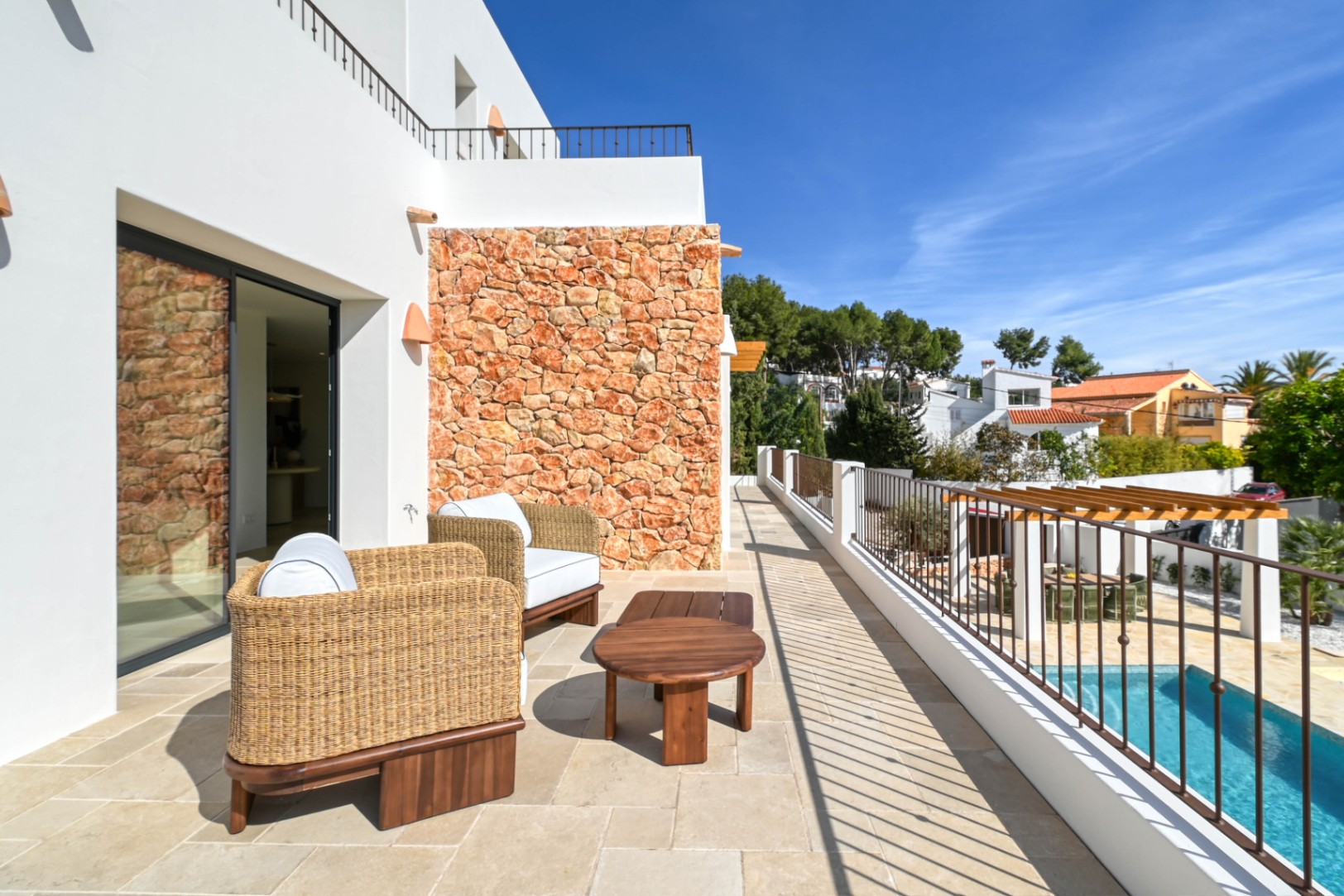 Obere Terrasse mit Natursteinfliesen, Korbsesseln und einer markanten Trockensteinmauer, mit Blick auf den Pool und den tiefer gelegenen Essbereich mit Pergola.