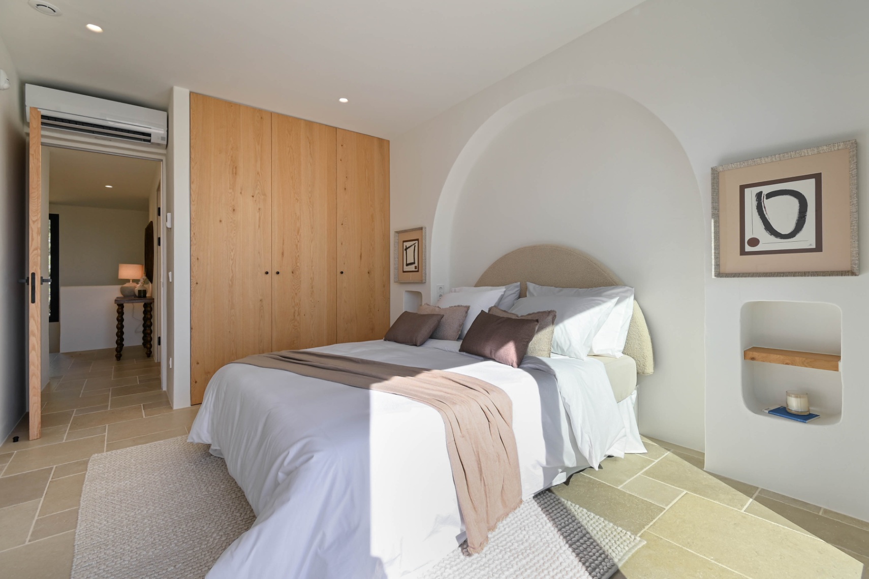 Schlafzimmer mit maßgefertigten Holzschränken, Natursteinboden und einer eleganten Rundbogennische. Ausgestattet mit Klimaanlage und dezenter Einbaubeleuchtung.