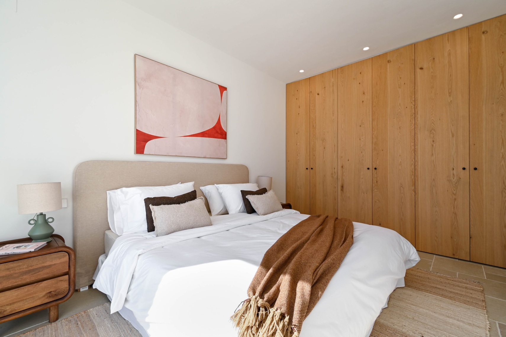 Schlafzimmer mit raumhohen Einbauschränken aus Naturholz, einem beigen Polsterkopfteil und Deckeneinbaustrahlern. Der Raum ist mit Fliesenboden und minimalistischem Dekor ausgestattet.