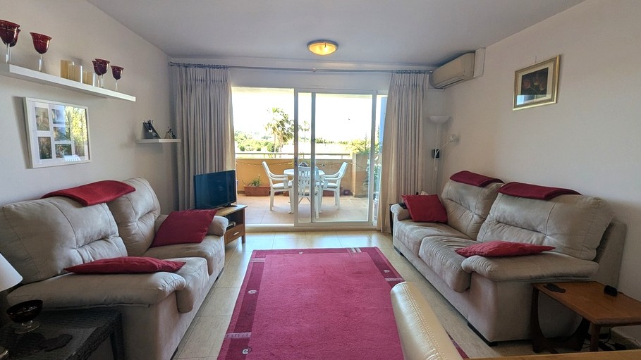 Helles Wohnzimmer mit neutralen Fliesenböden, zwei Sofas mit roten Akzenten und Glasschiebetüren, die auf eine möblierte Terrasse mit Blick ins Grüne führen.