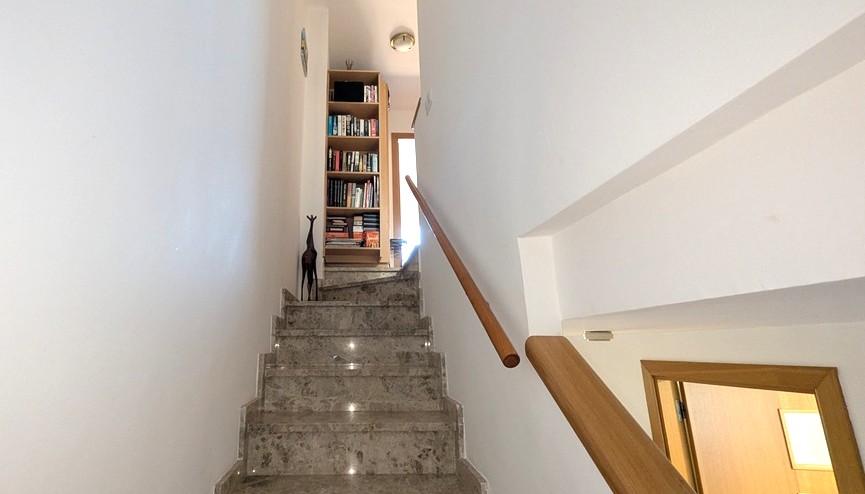 Innentreppe aus Marmor mit Holzhandlauf zum Obergeschoss, ausgestattet mit einem eingebauten Bücherregal und hellen, weißen Wänden.