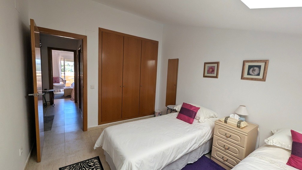 Schlafzimmer mit zwei Einzelbetten, hölzernen Einbauschränken und Fliesenboden. Die offene Tür gibt den Blick frei auf den Flur und ein Zimmer mit Terrassenzugang.