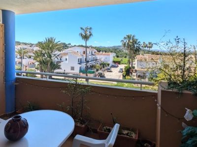 Balkon mit Tisch und Stuhl, mit freiem Blick auf ein Wohngebiet mit weißen Häusern, Palmen und grünen Hügeln unter strahlend blauem Himmel.