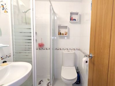 Das moderne Duschbad verfügt über weiß geflieste Wände mit Zierleiste, eine Duschkabine aus Glas mit Milchglasstreifen, ein Wandwaschbecken und eine Toilette. Zwei quadratische Wandregale bieten Stauraum.