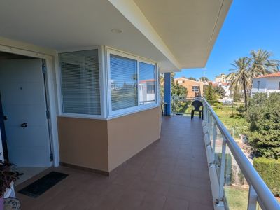 Geräumige private Terrasse mit braunem Fliesenboden, Glasgeländer und teilweisem Fensterbereich. Bietet Blick auf Palmen und die Wohnlandschaft.