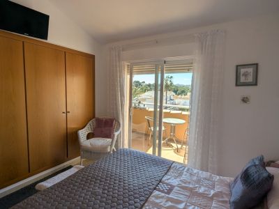 Schlafzimmer mit integriertem Holzschrank und Zugang zu einem privaten Balkon, der von seiner kleinen Sitzecke aus Blicke auf Dächer, Palmen und ferne Hügel bietet.