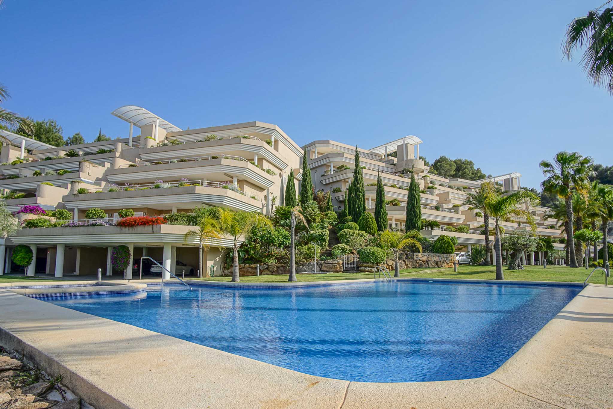 Außenansicht der Anlage Las Mimosas mit großem Gemeinschaftspool und terrassierter Architektur, eingebettet in gepflegte mediterrane Gartenanlagen mit Palmen.