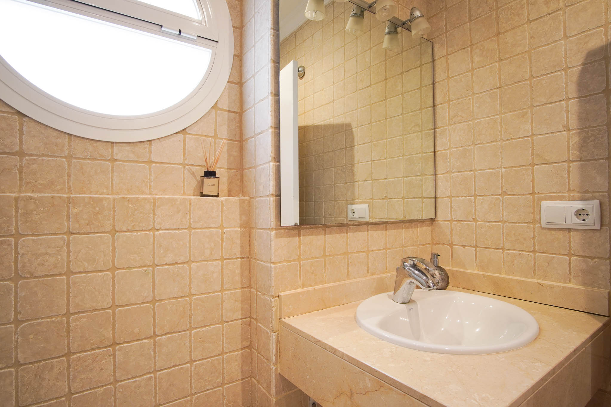 Badezimmer mit getrommelten Marmorfliesen, Steinarbeitsplatte mit Einbauspülbecken und einem markanten runden Fenster für natürliches Licht.