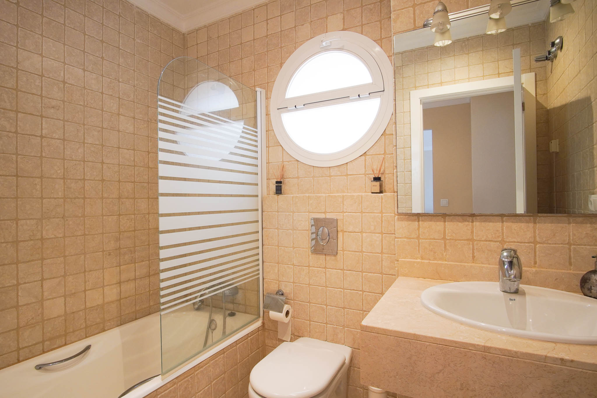 Badezimmer mit Marmoroberflächen, Badewanne mit Glasabtrennung und einem markanten runden Fenster für natürliches Licht.