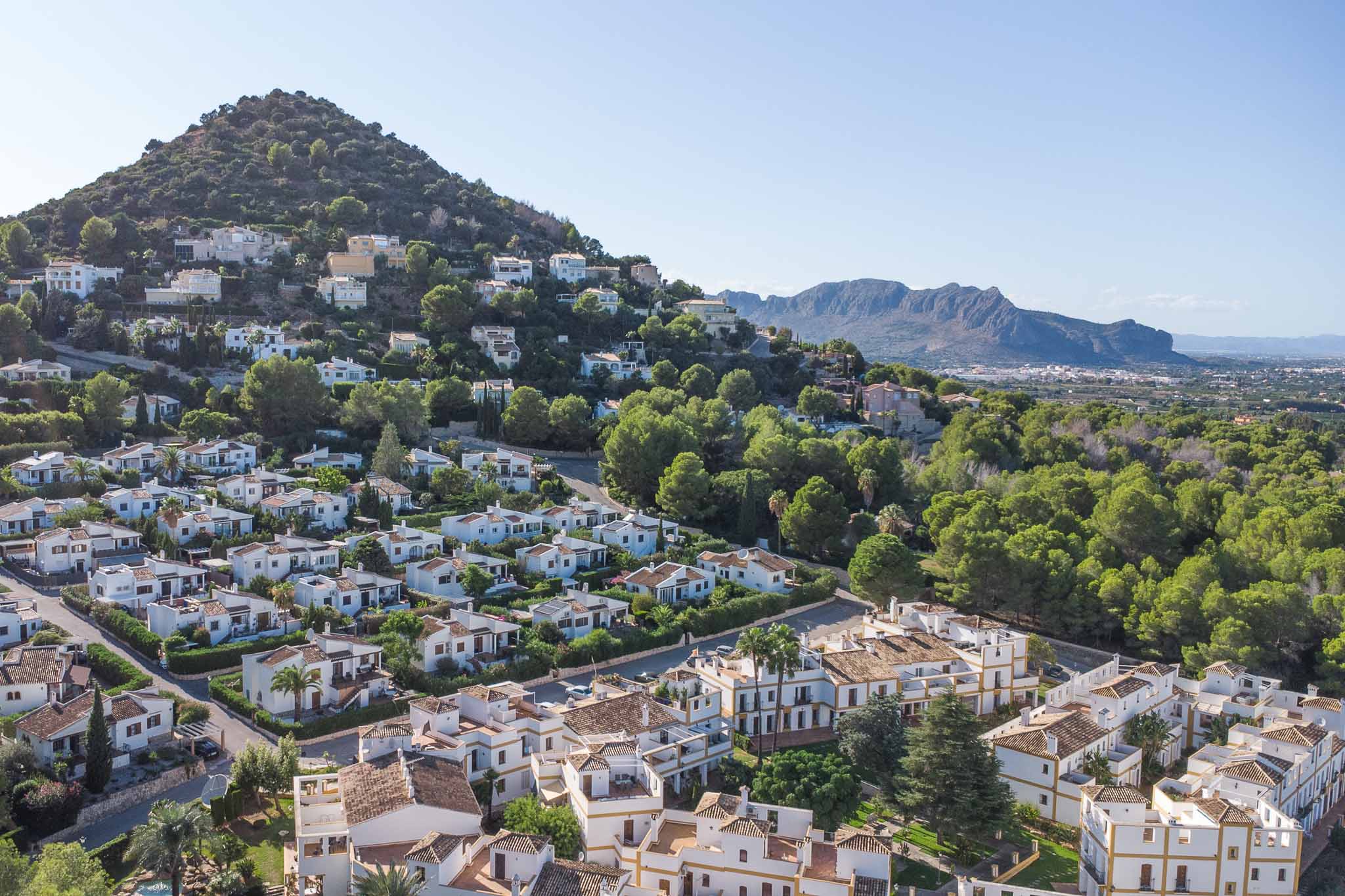 Erhöhter Panoramablick auf das Wohngebiet La Sella mit mediterraner Architektur, eingebettet in einen Pinienwald vor einer markanten Bergkulisse.