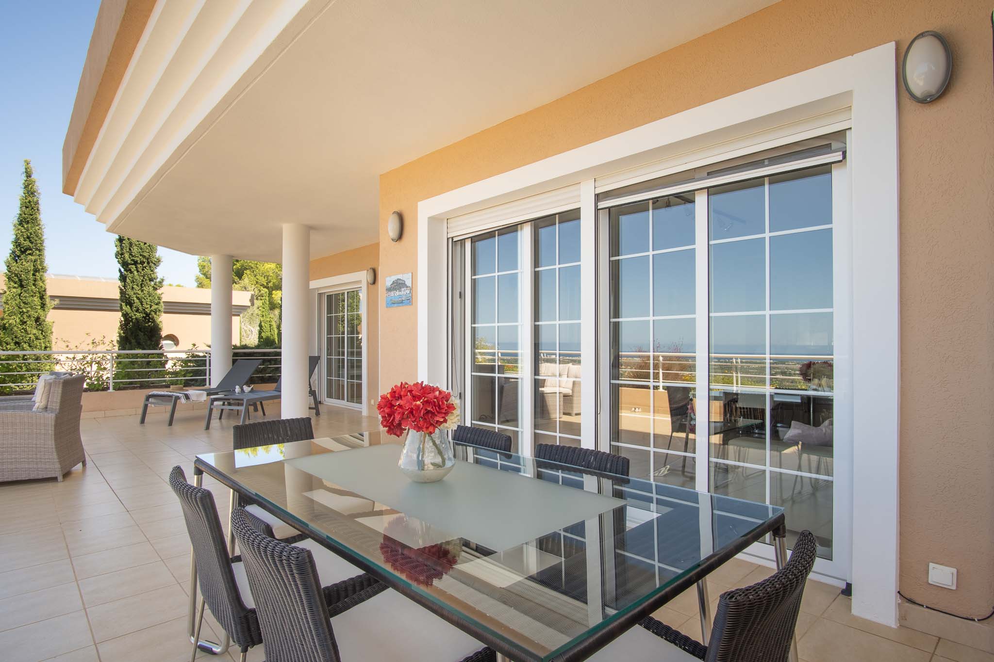 Großzügige überdachte Terrasse mit Essbereich im Freien, Glastisch und Korbstühlen, verbunden durch große Glasschiebetüren mit Blick auf das Meer.