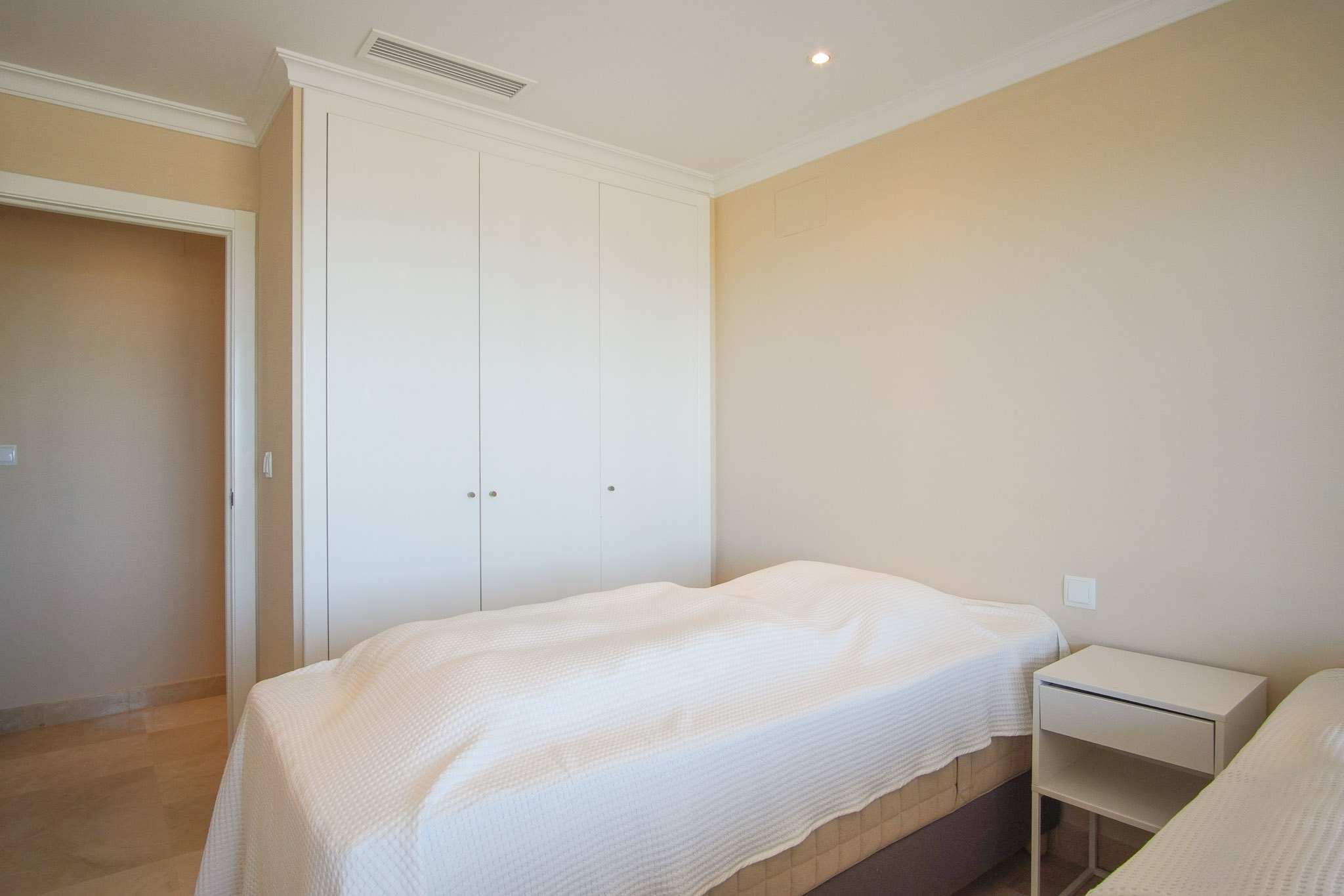 Helles Schlafzimmer mit weißen Einbauschränken, Marmorboden und integrierter Klimaanlage. Cremefarbene Wände und klassische Deckenstuckleisten.