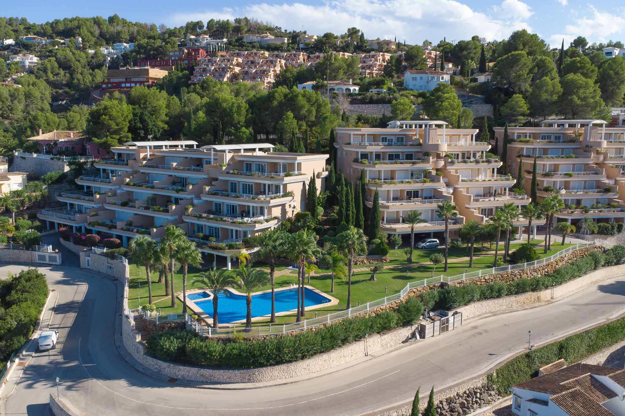 Luftaufnahme der Anlage Las Mimosas in La Sella mit terrassierter Architektur, Gemeinschaftspool mit Palmen und gepflegten Grünflächen.