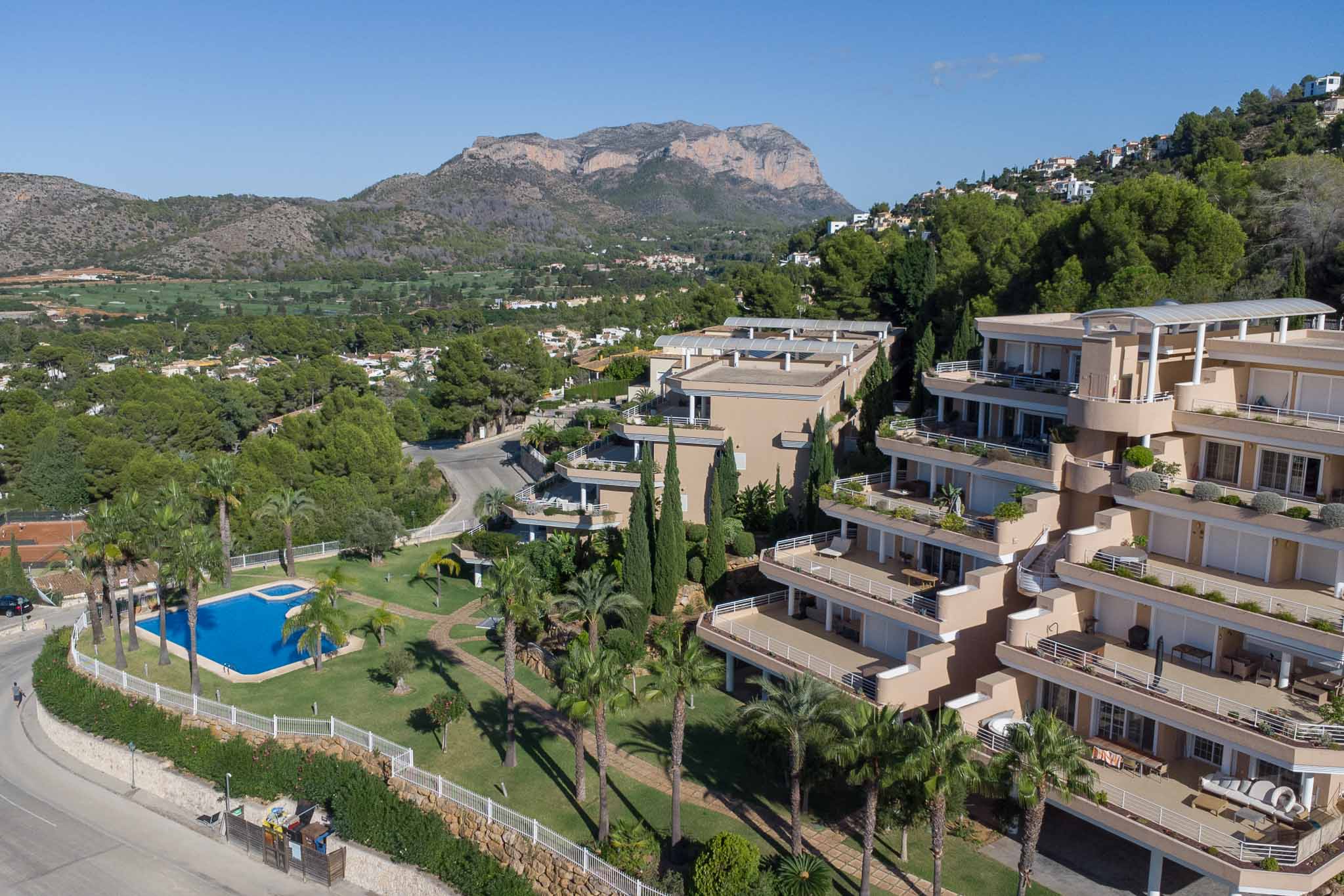 Luftaufnahme der Residenz Las Mimosas mit Gemeinschaftspool, Palmengärten und terrassierten Apartments mit Blick auf den Golfplatz und das Montgó-Massiv.