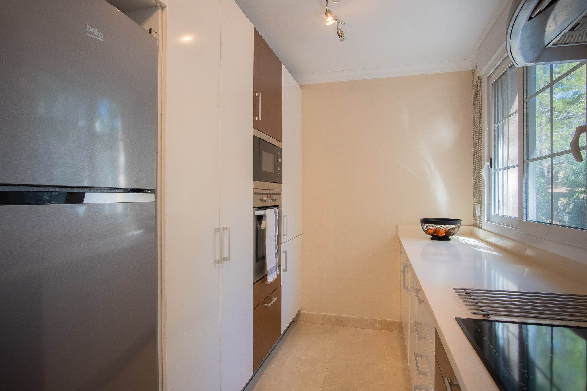Moderne Küche mit weiß- und holzfarbenen Schränken, integrierten Edelstahlgeräten und weißer Arbeitsplatte. Ein Seitenfenster sorgt für natürliches Licht am Induktionskochfeld.