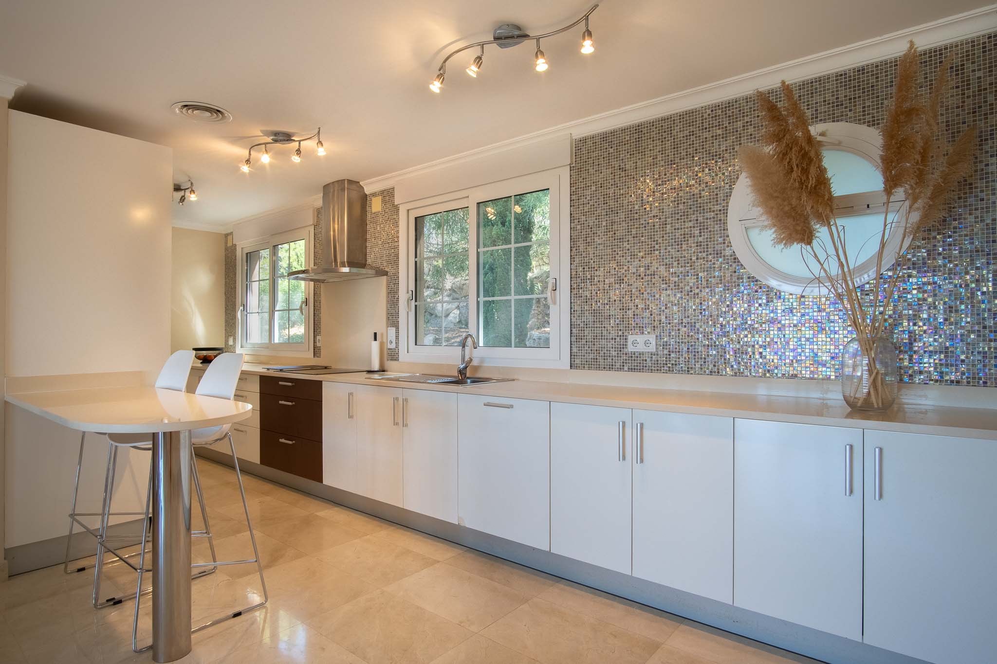 Moderne Küche mit weißen Schränken, heller Arbeitsplatte und schillerndem Mosaik-Fliesenspiegel. Mit Frühstückstisch, Marmorboden und Fenstern für reichlich natürliches Licht.