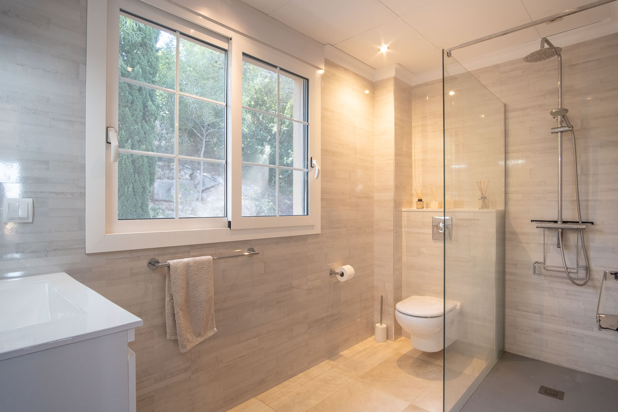 Modernes Badezimmer mit begehbarer Dusche, Glastrennwand und sandfarbenen Fliesen. Ausgestattet mit wandhängendem WC, Einbaustrahlern und Fenster mit Blick in den Garten.