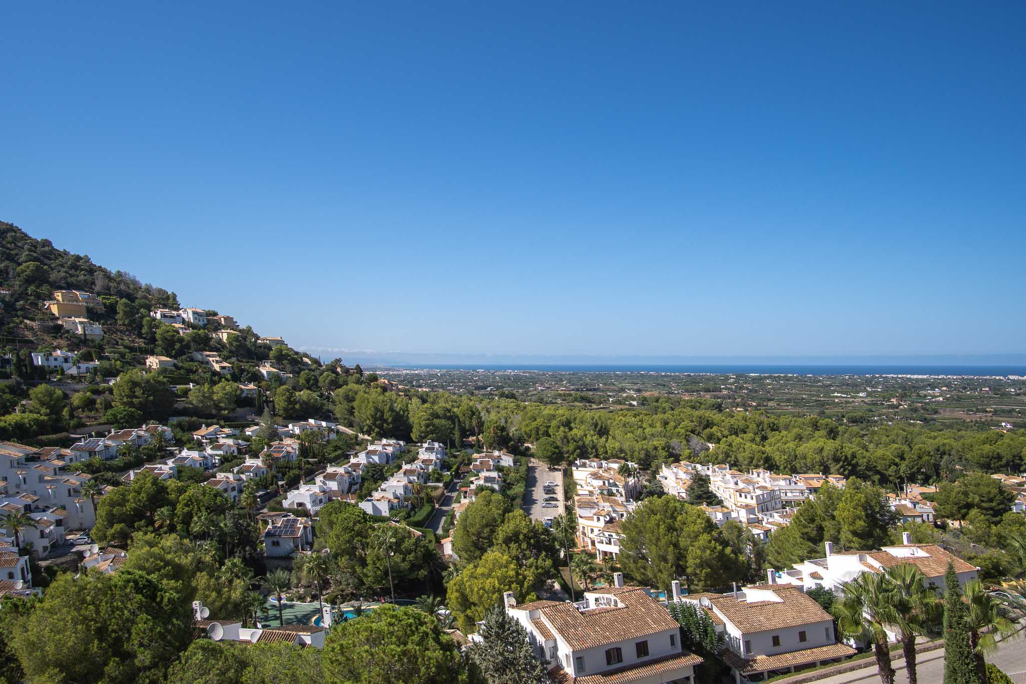 Panoramablick von La Sella auf das Mittelmeer, der die in die üppige grüne Umgebung integrierte Wohnarchitektur unter klarem Himmel zeigt.