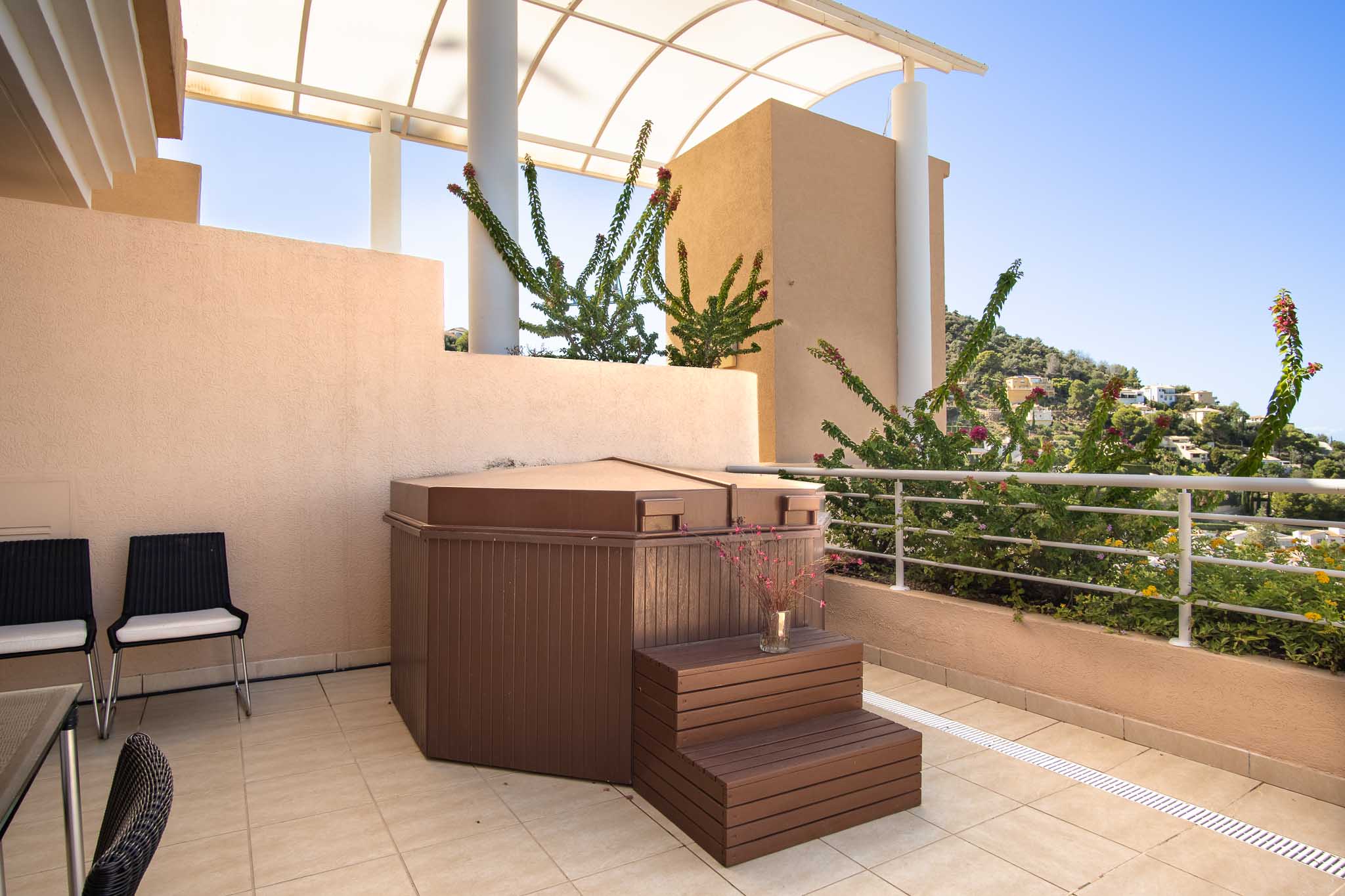 Private Terrasse mit abgedecktem Whirlpool und Holzstufen unter einer gewölbten Pergola, mit Blick auf die Hügel von La Sella und mediterrane Pflanzen.