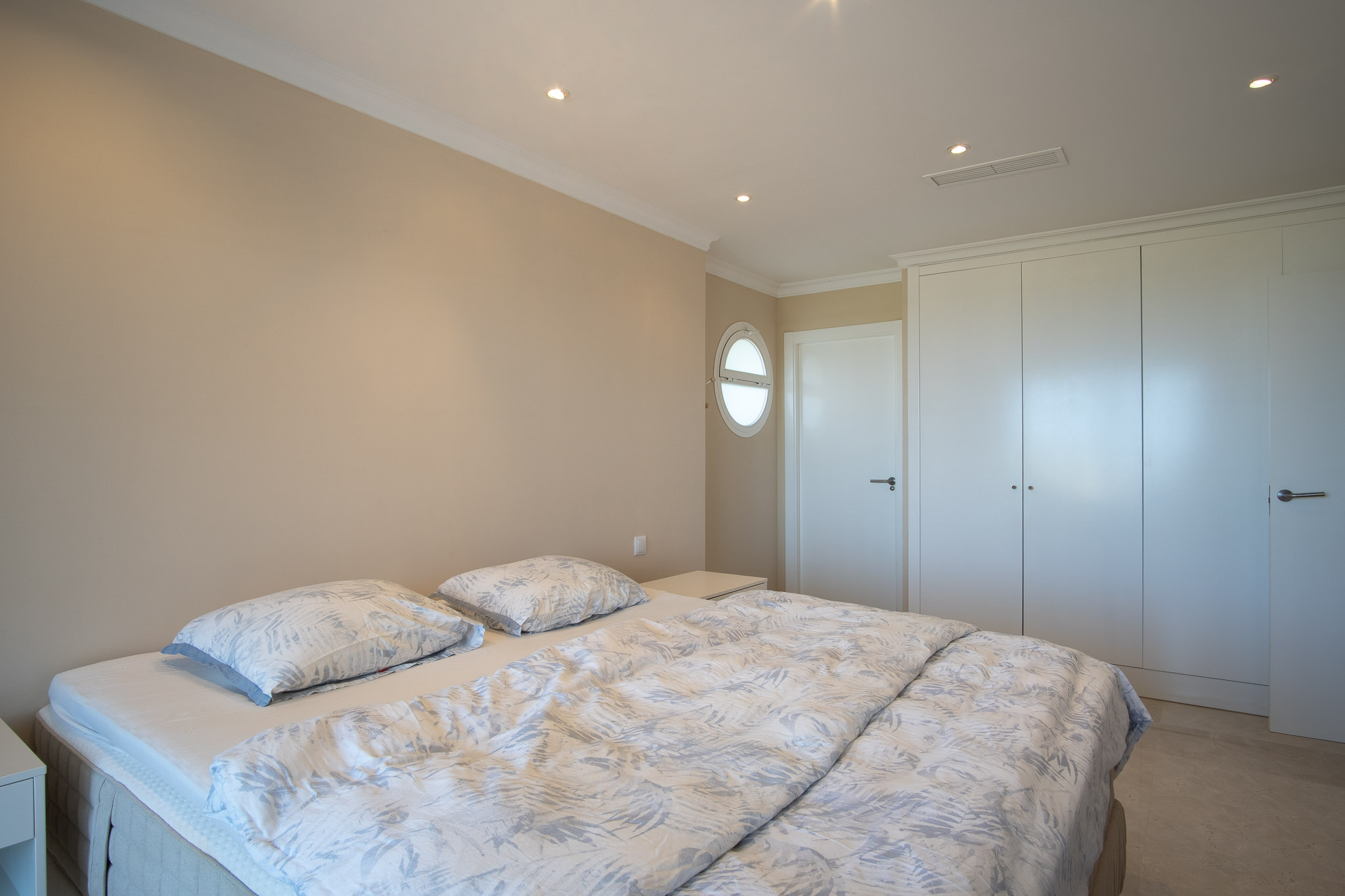 Schlafzimmer mit Doppelbett, weißen Einbauschränken und Marmorboden. Ausgestattet mit Decken-Einbaustrahlern und einem markanten Bullaugenfenster.
