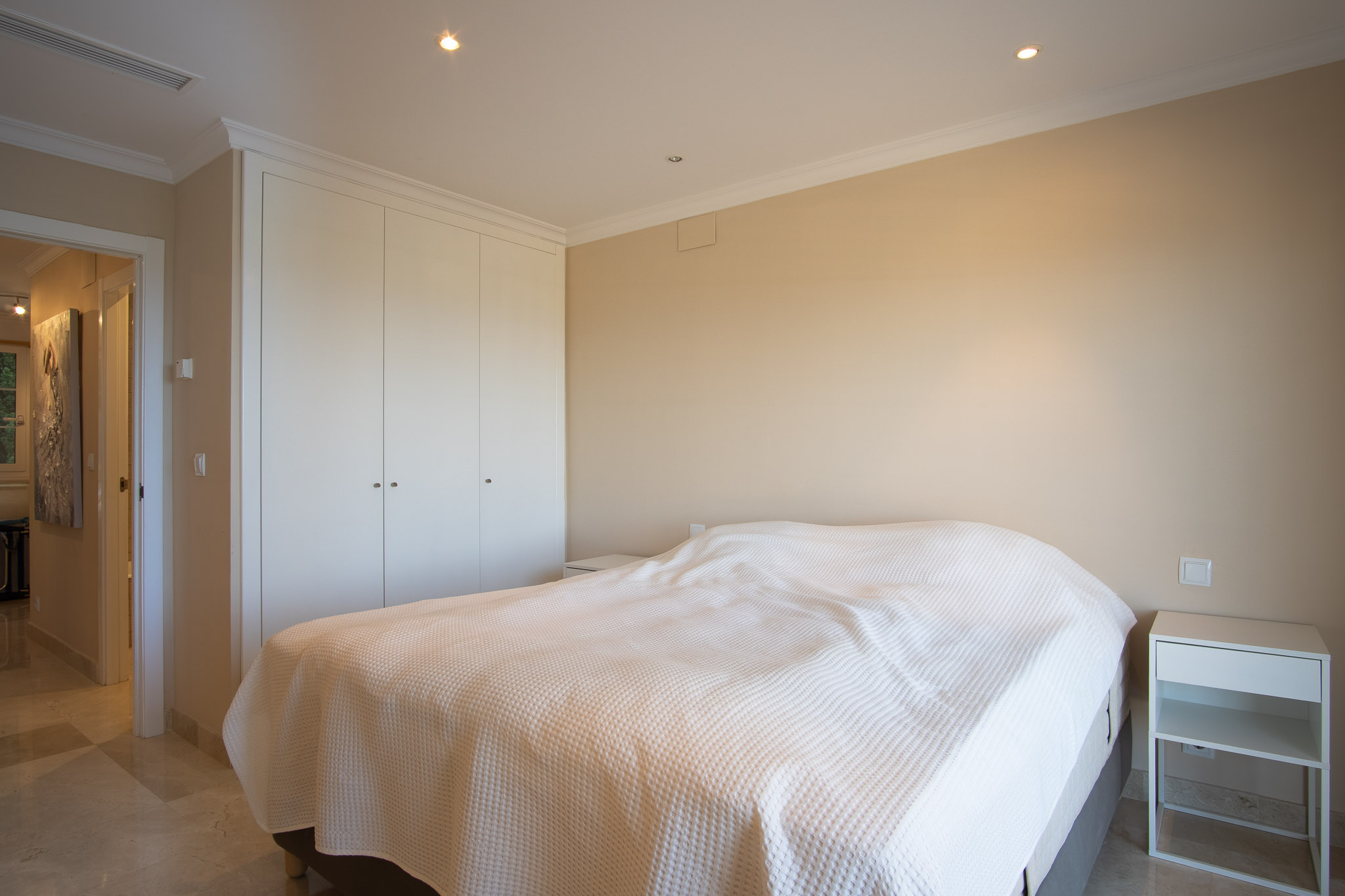 Schlafzimmer mit Marmorboden, deckenhohen weißen Einbauschränken und Deckeneinbaustrahlern, das durch sein schlichtes und funktionales Design besticht.
