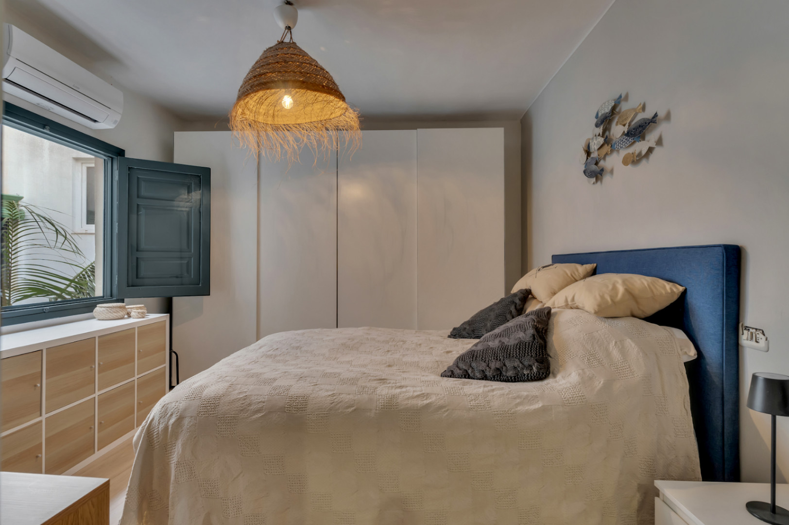 Helles Schlafzimmer mit blauem Kopfteil, Korblampe und Fenster mit Fensterläden.