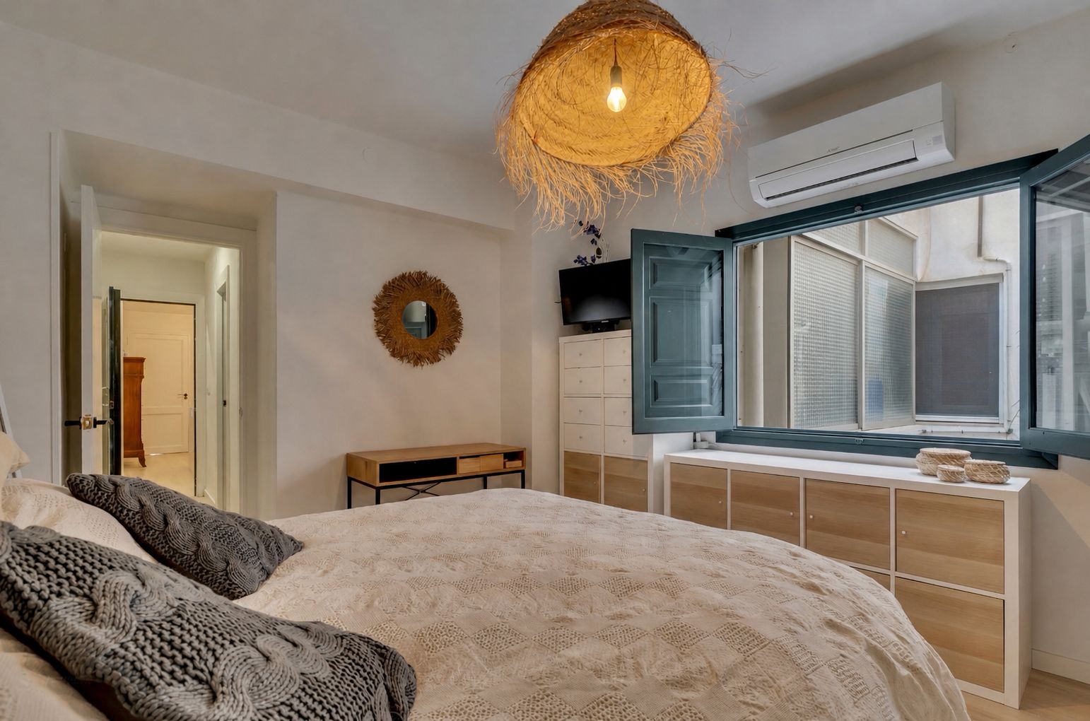 Helles Schlafzimmer mit Doppelbett, blauem Holzfensterrahmen und hängender Lampe aus Korbgeflecht.