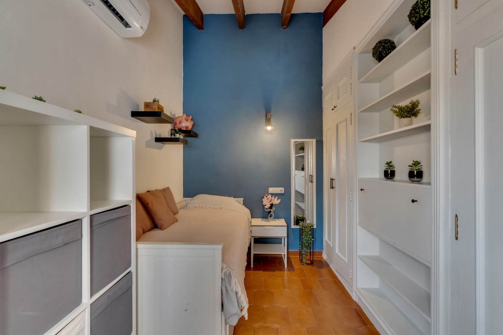 Schlafzimmer mit blauer Akzentwand, weißen Regalen und Terrakottafliesenboden. Sichtbare Holzbalken an der Decke.