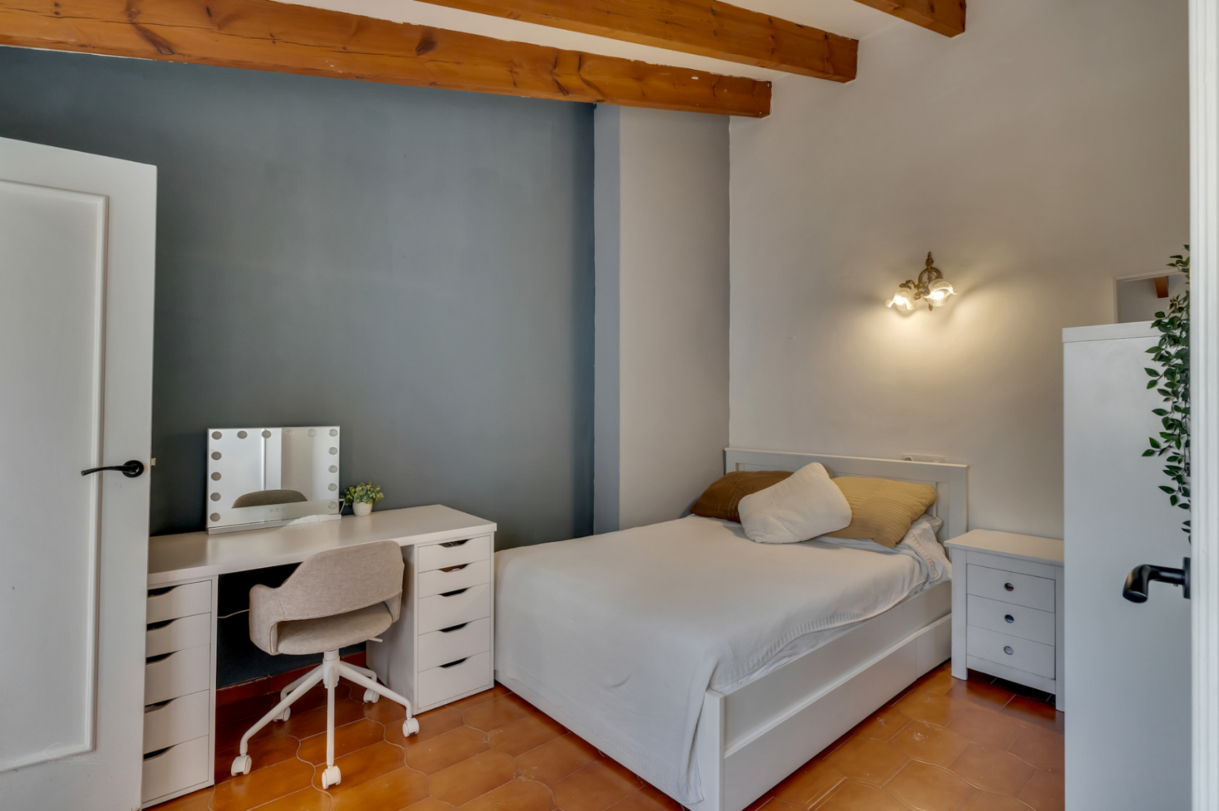 Schlafzimmer mit Terrakottaboden, blauer Akzentwand und weißem Schreibtisch mit beleuchtetem Spiegel.