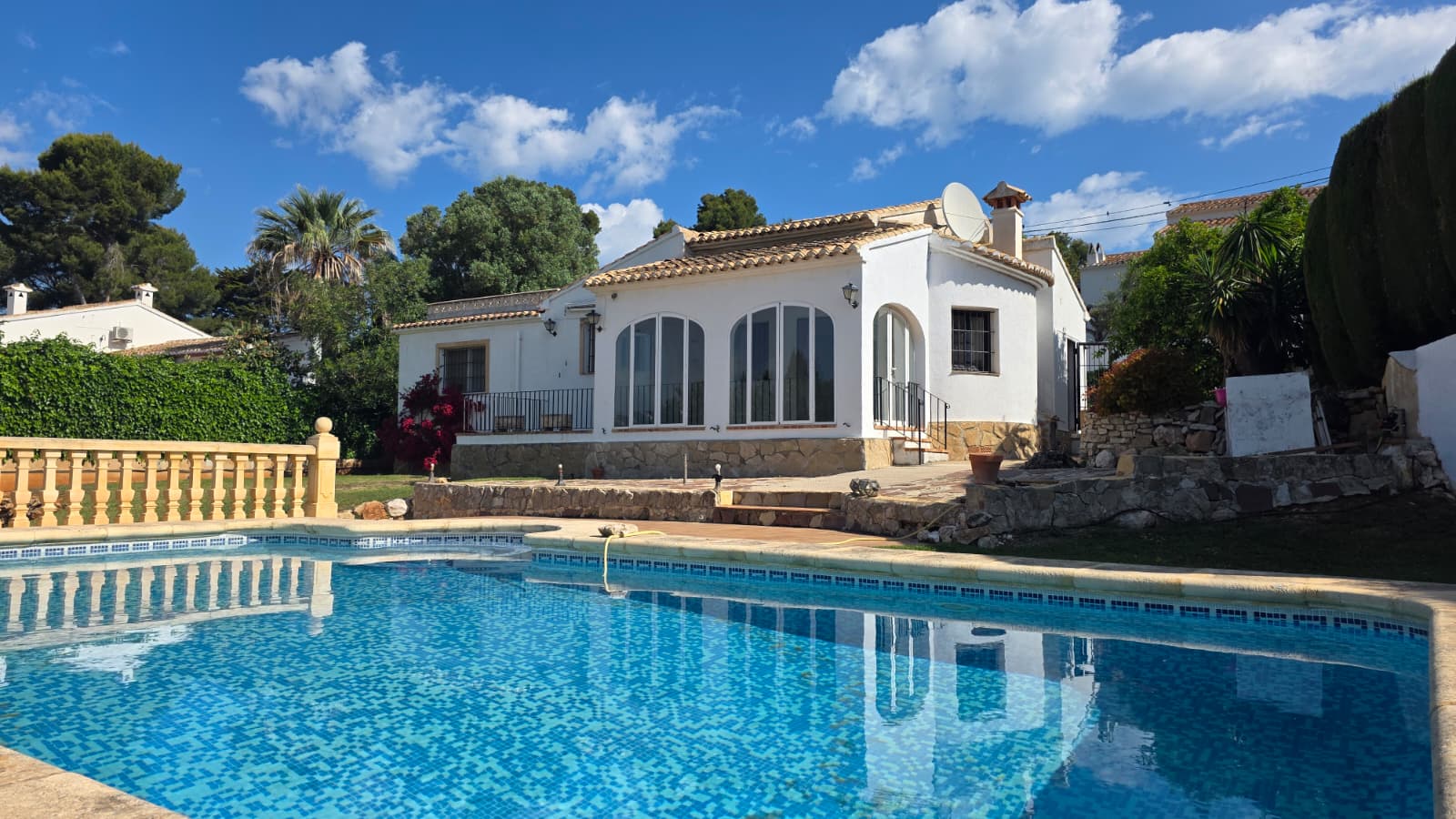 Außenansicht des privaten Pools und der weißen Villa mit traditionellen Rundbogenfenstern und Natursteinsockel in der Urbanisation El Tosalet, Jávea.