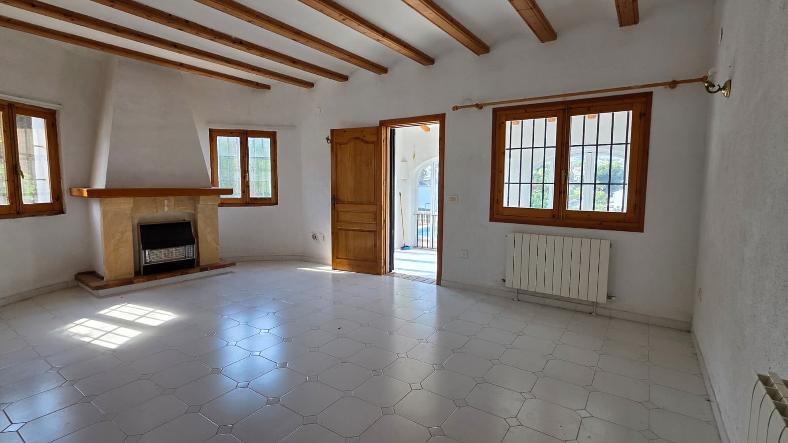 Wohnzimmer mit Holzbalkendecke, Tosca-Steinkamin und weißem Fliesenboden. Große Holzfenster und Zugang zur Terrasse sorgen für helles Tageslicht.