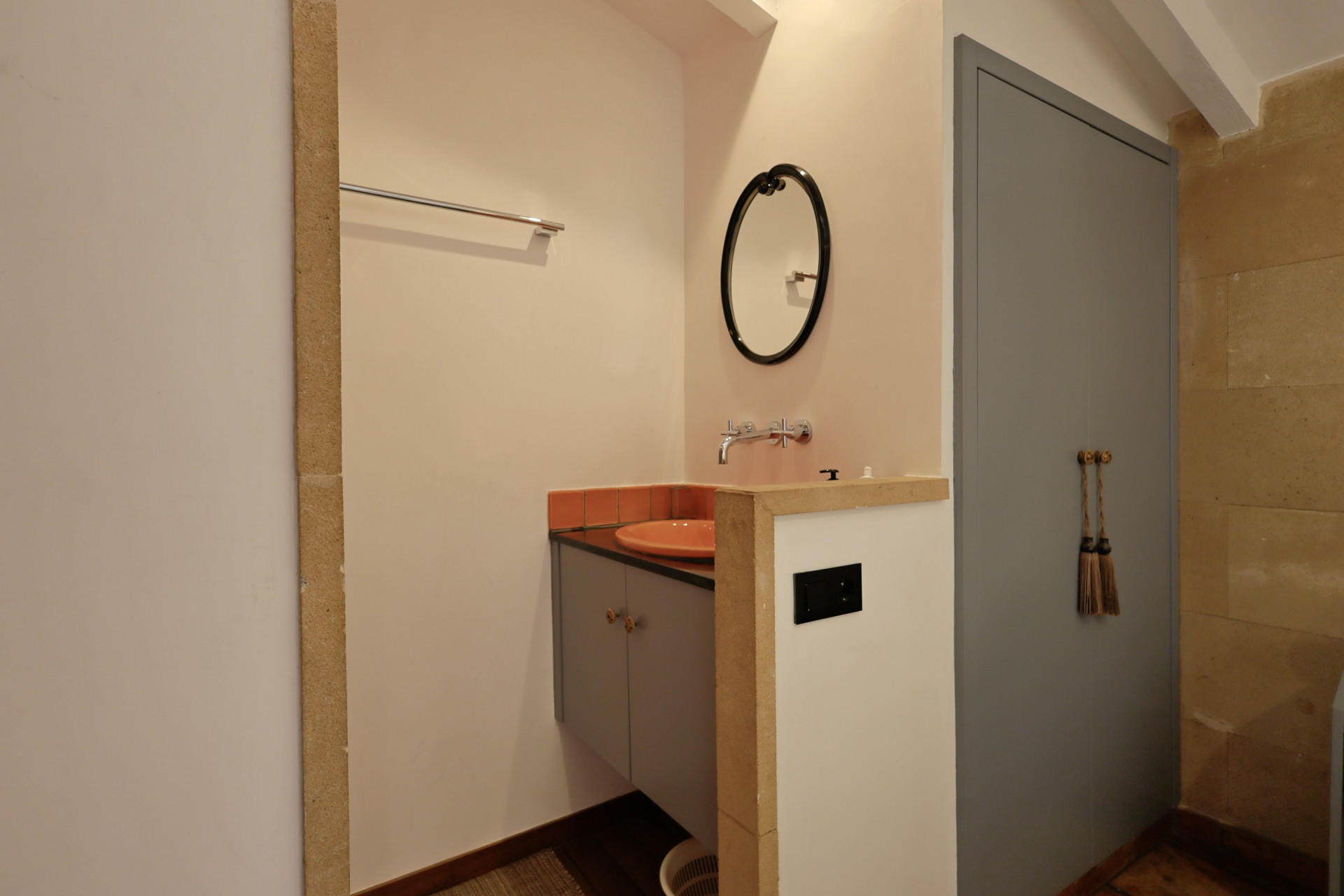 Badezimmerdetail mit orangefarbenem Keramikwaschbecken, ovalem Spiegel und grauer Tür mit dekorativen Quasten.