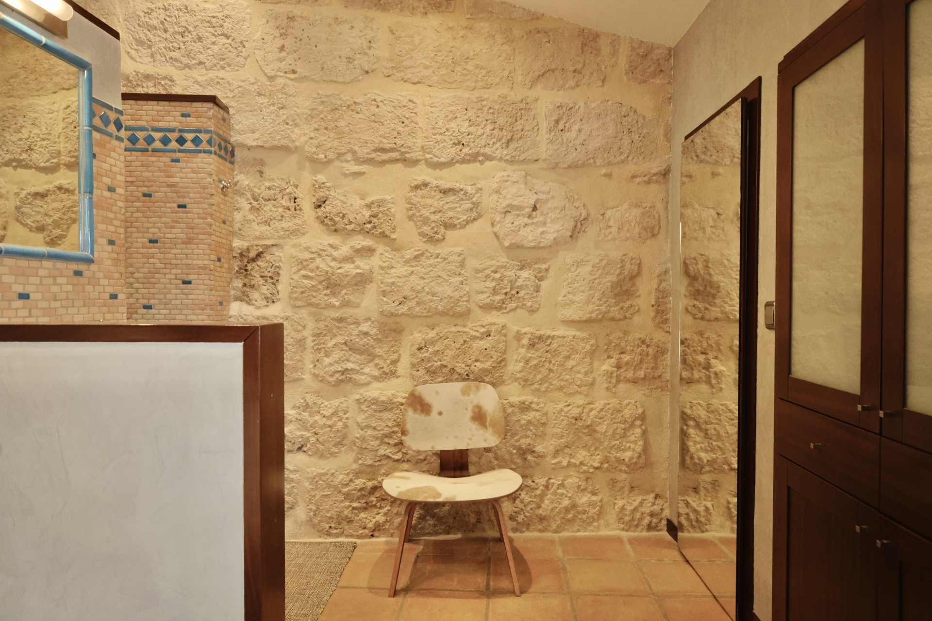 Badezimmerdetail mit rustikaler Steinwand, blau gerahmtem Spiegel und Designerstuhl mit Kuhfellpolsterung.