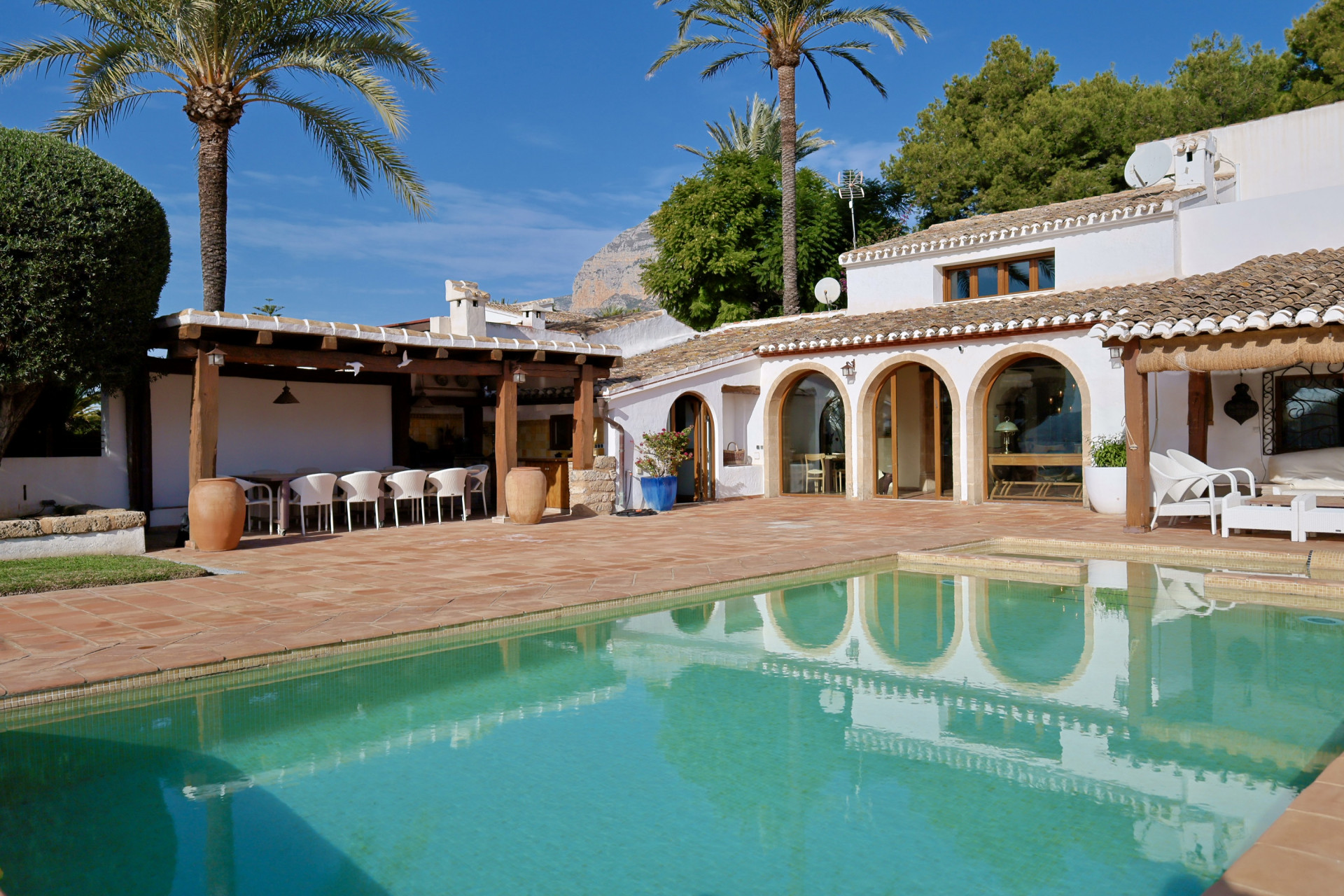 Blick auf den türkisfarbenen Pool, der die Bogenarchitektur und die Pergola mit Essbereich im Freien widerspiegelt.