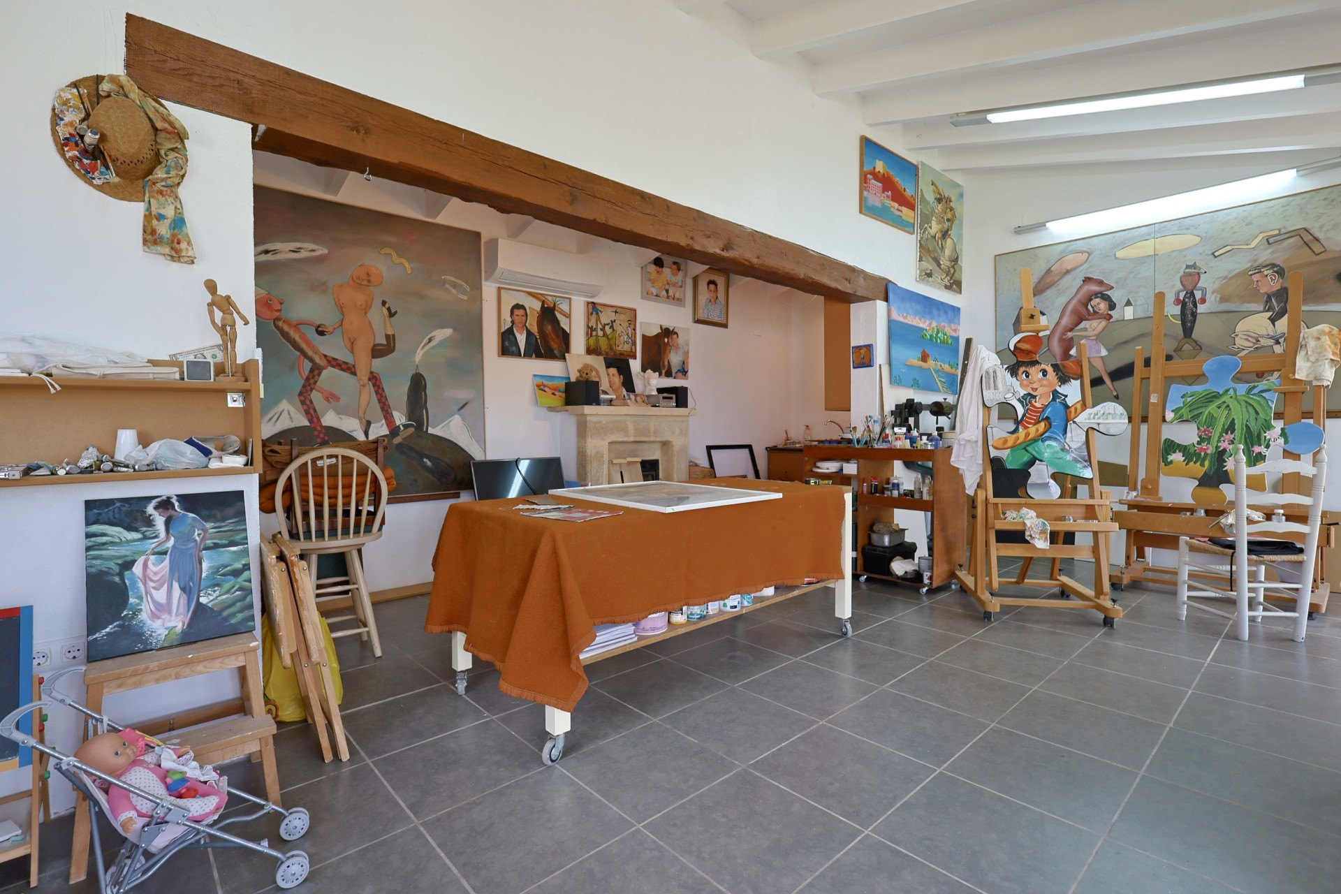 Geräumiges Atelier mit grau gefliesten Böden, einem orangefarbenen Tuch bedeckten Tisch und Staffeleien mit Gemälden.
