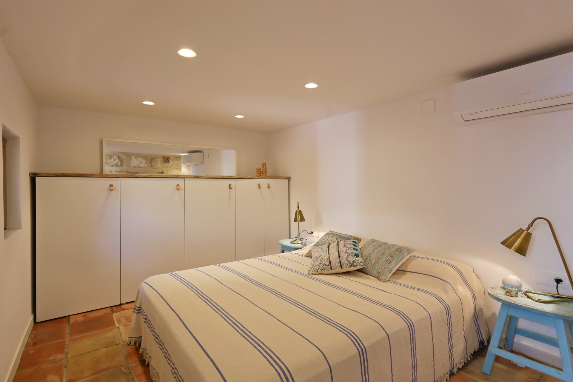 Helles Schlafzimmer mit Terrakottaboden, gestreifter Bettdecke und weißem Einbauschrank mit Holzdetails.