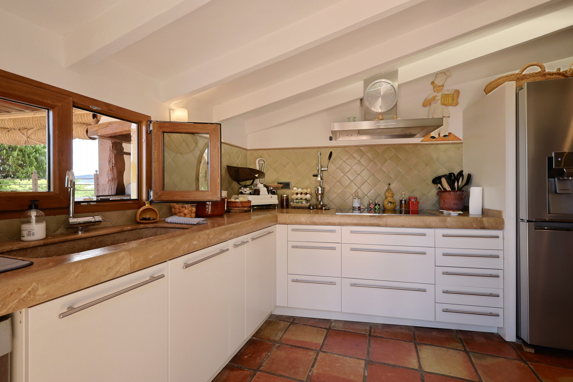 Rustikale Küche mit Steinarbeitsplatte, weißen Schränken und Terrakottaboden. Steinspüle mit Holzfenster.