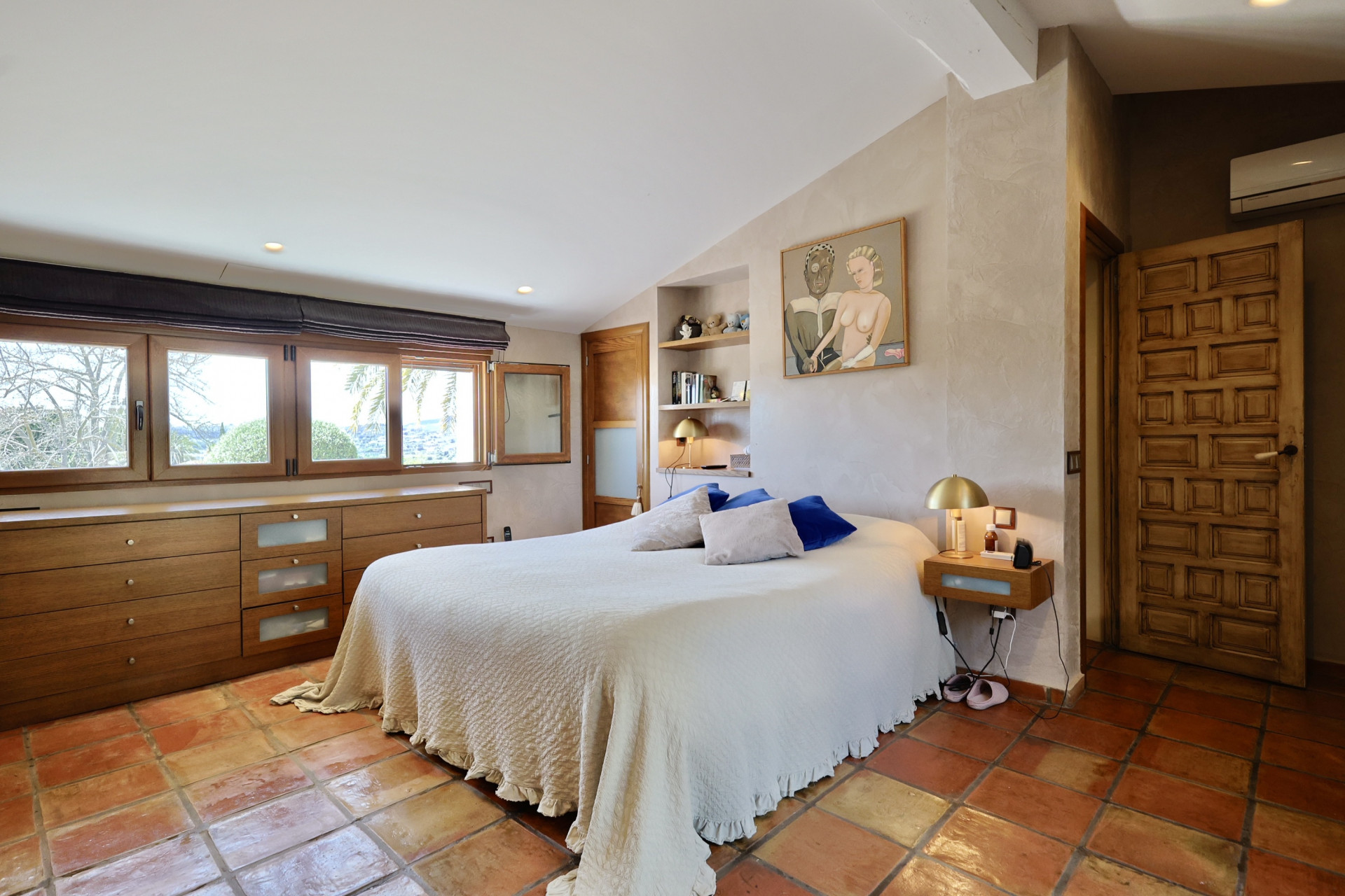 Schlafzimmer mit Terrakottafliesen, Doppelbett mit weißer Bettwäsche und Holzkopfteil mit Fenstern.