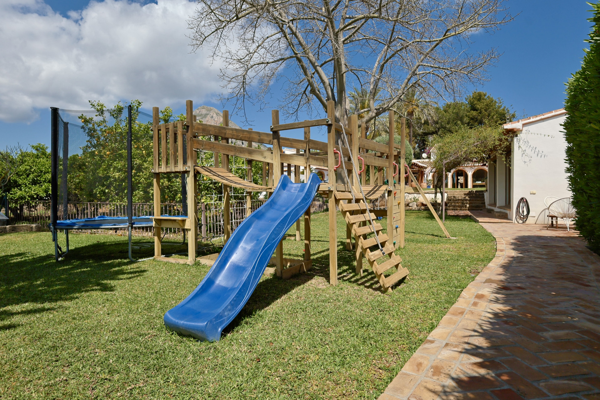 Spielbereich mit Holzstruktur, blauer Rutsche und Trampolin im Garten.