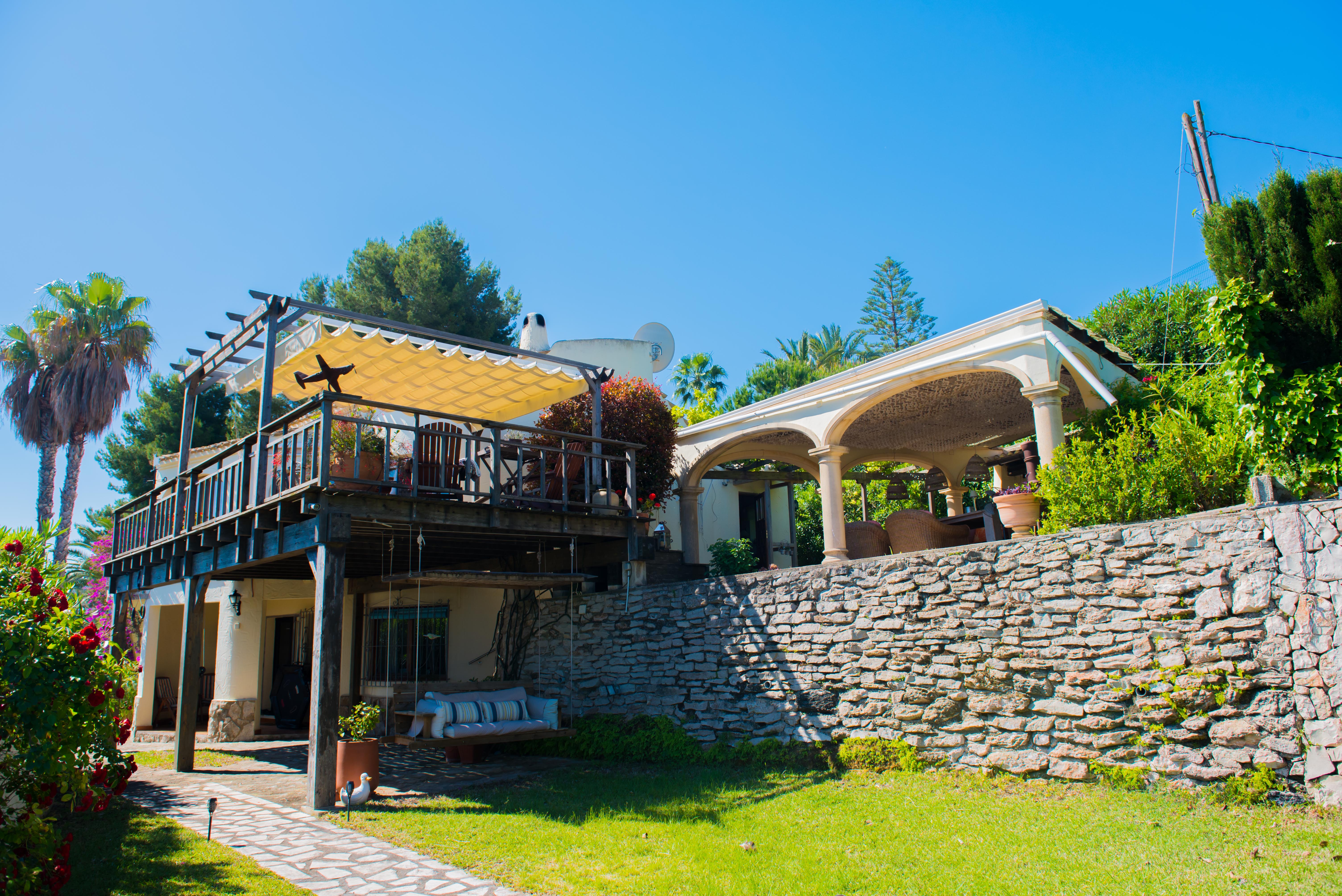 Außenansicht der Villa mit überdachter Holzterrasse und Markise, Garten mit Steinmauer und Ruhebereich.