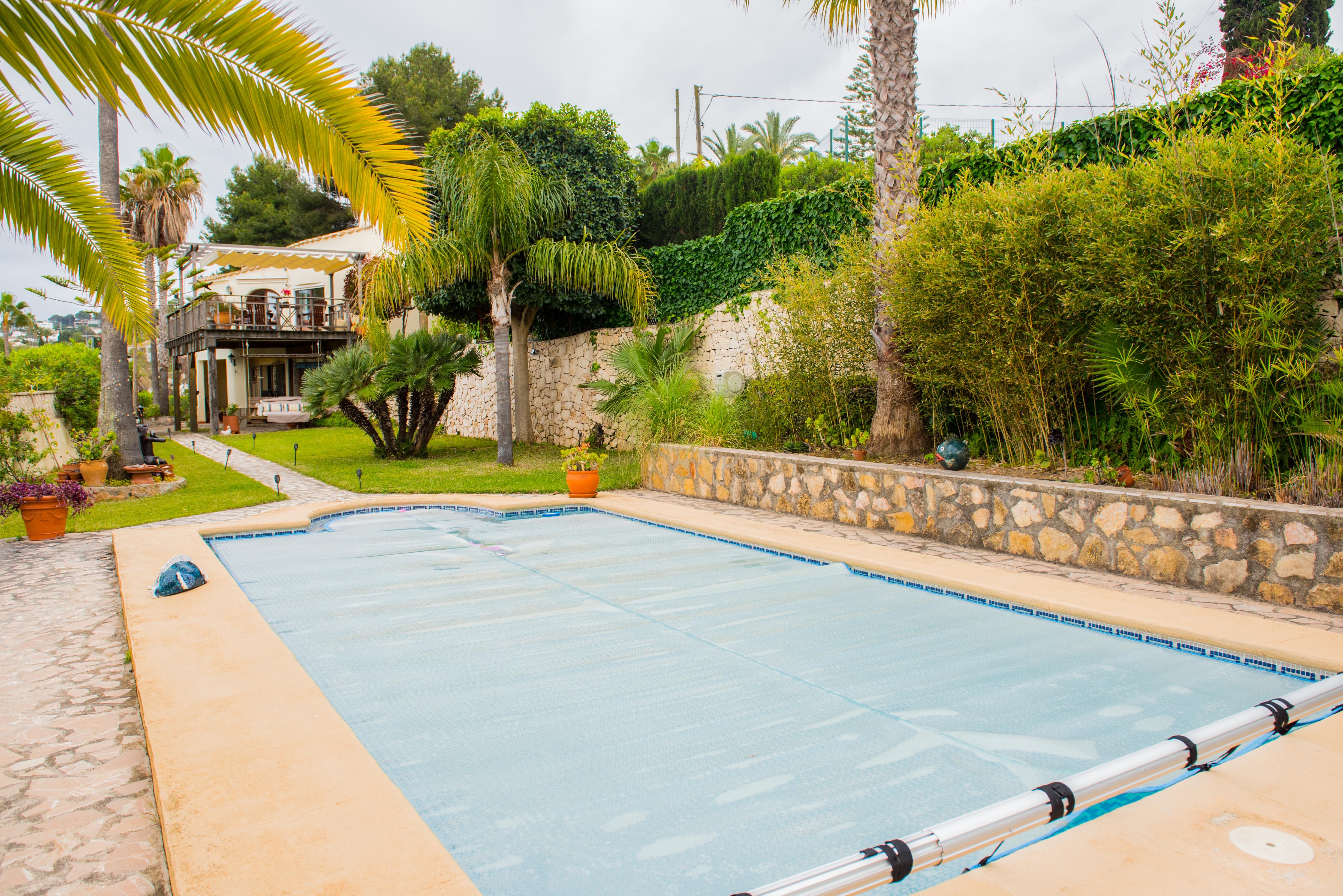 Außenpool mit blauer Thermoabdeckung und Aufrollvorrichtung, eingebettet in einen Garten mit Natursteinmauern, Palmen und üppiger mediterraner Bepflanzung.