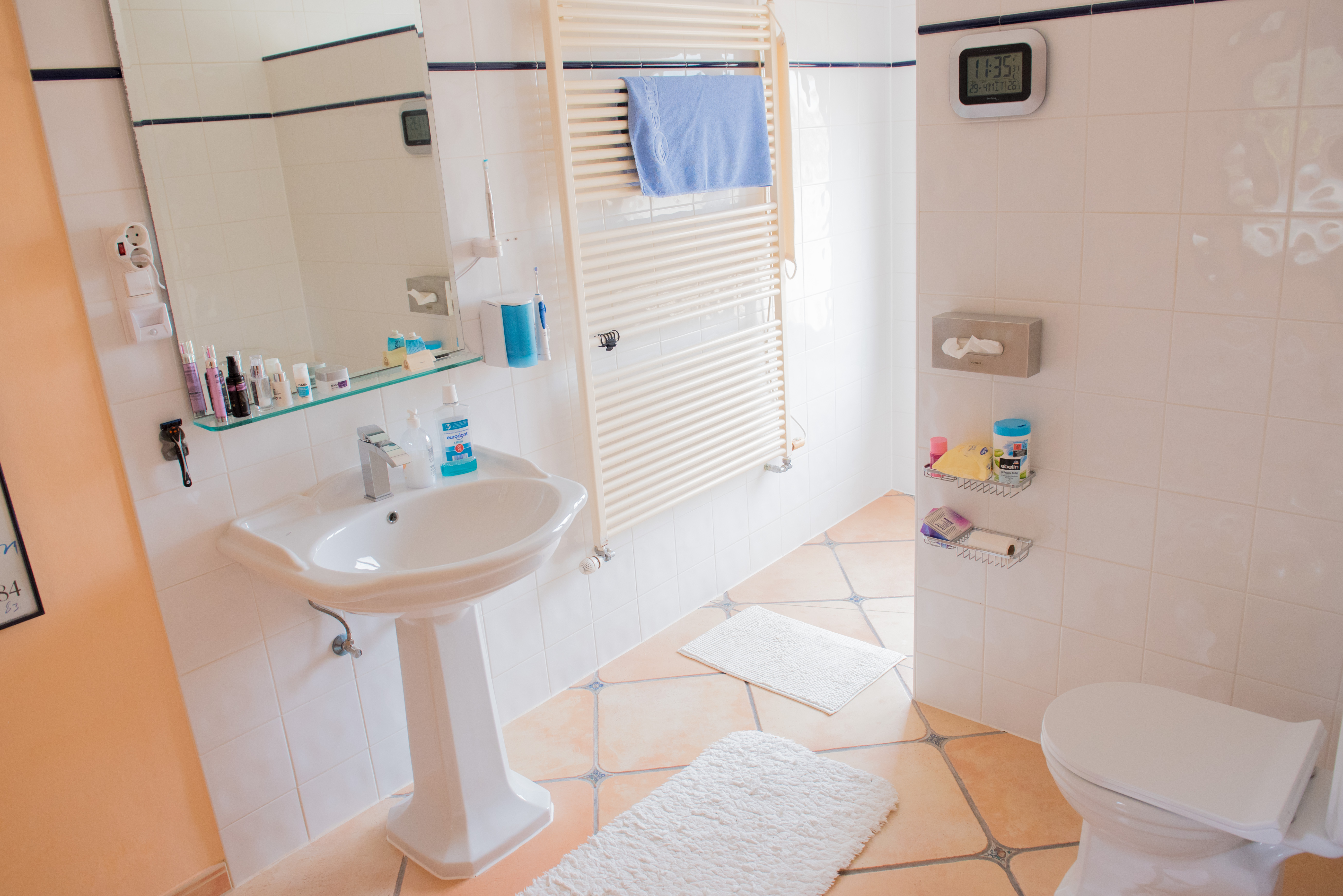 Badezimmer mit Standwaschbecken, Terrakotta-Fliesenboden mit Dekorelementen, Handtuchheizkörper und weißen Wandfliesen mit blauer Bordüre.
