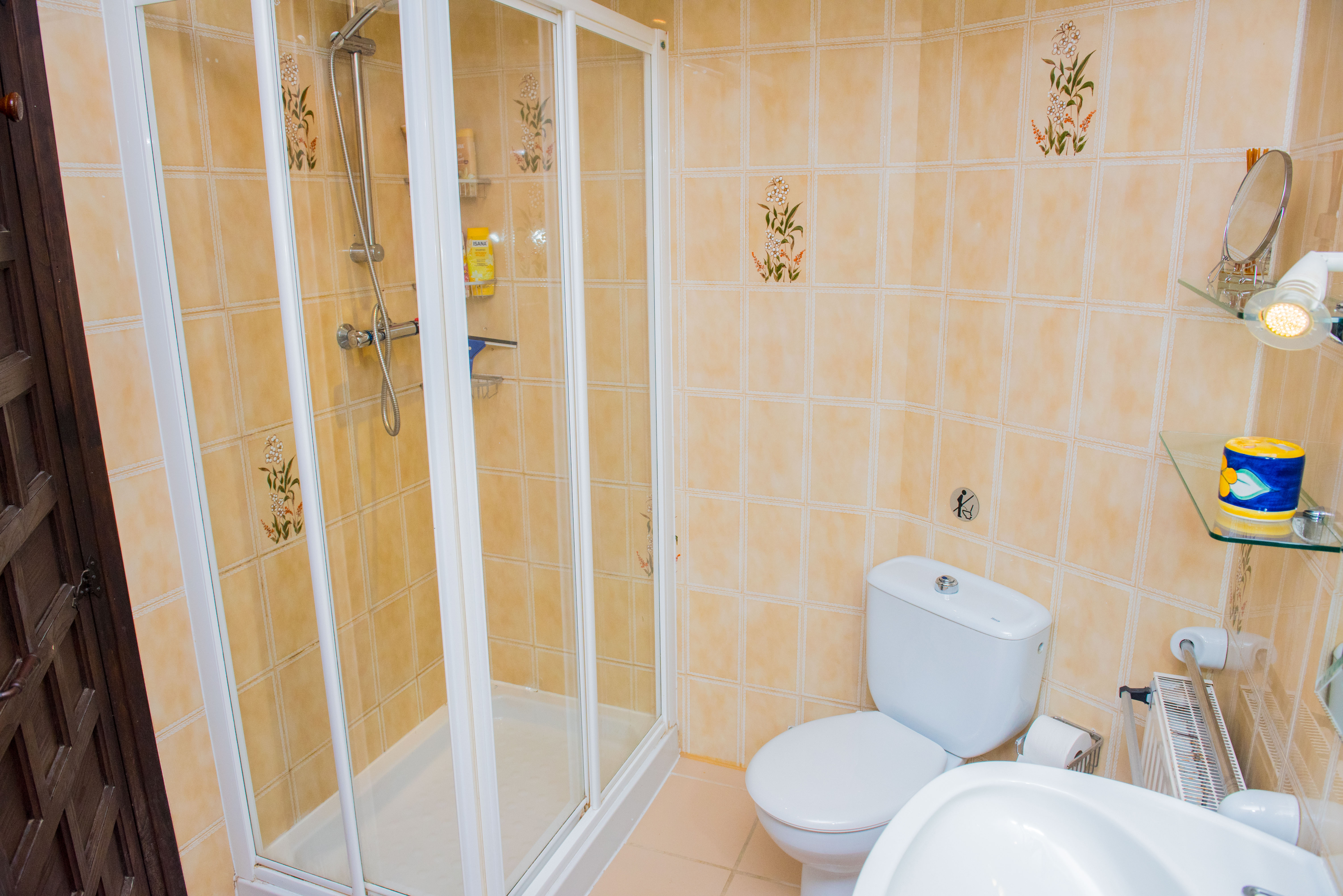 Badezimmer mit weiß gerahmter Glasduschkabine, beigen Wandfliesen mit floralen Dekoren und weißen Keramik-Sanitärobjekten.