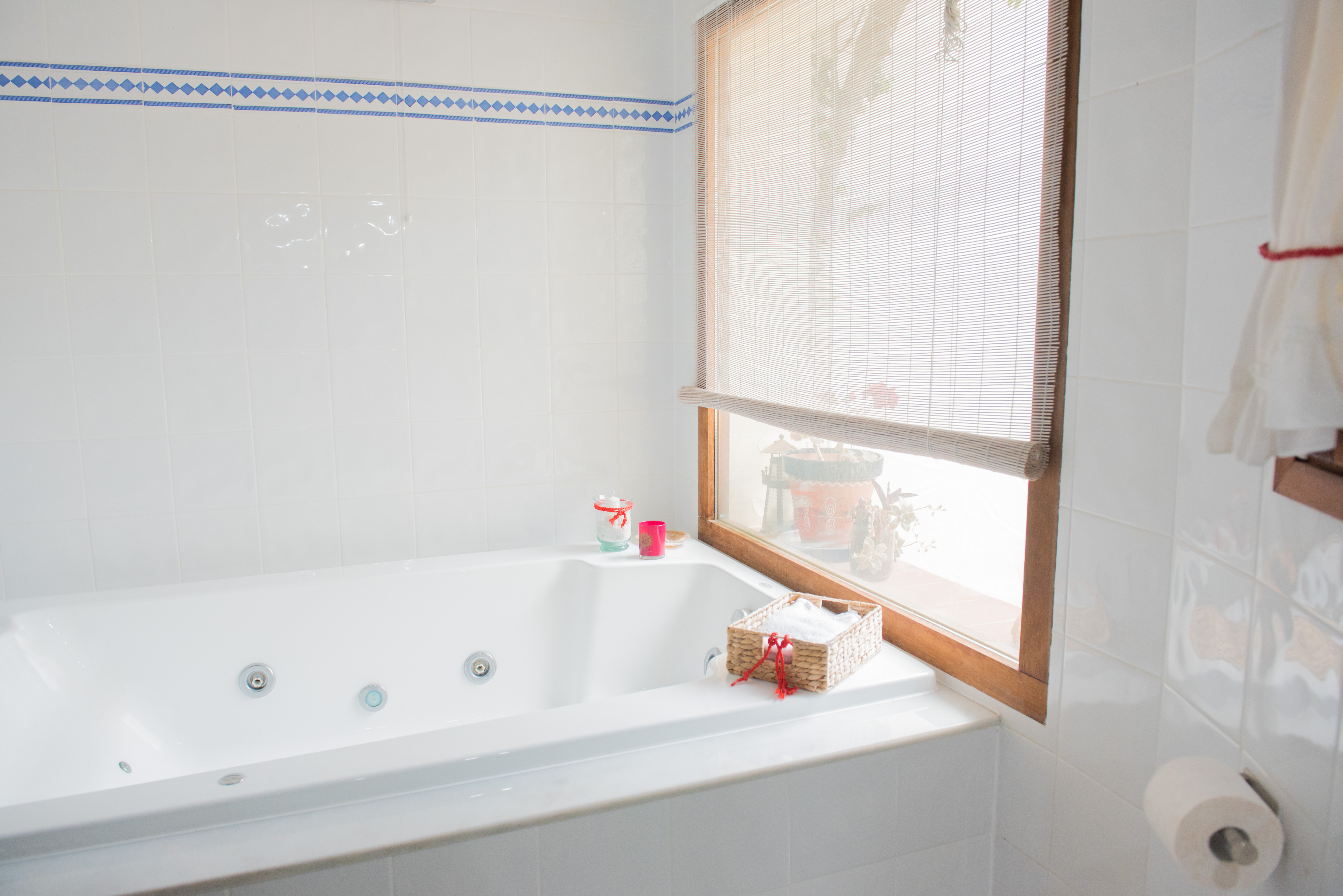 Badezimmer mit Whirlpool-Badewanne, weißen Wandfliesen mit blauer Bordüre und einem Holzfenster mit Bambusrollo für helles Tageslicht.