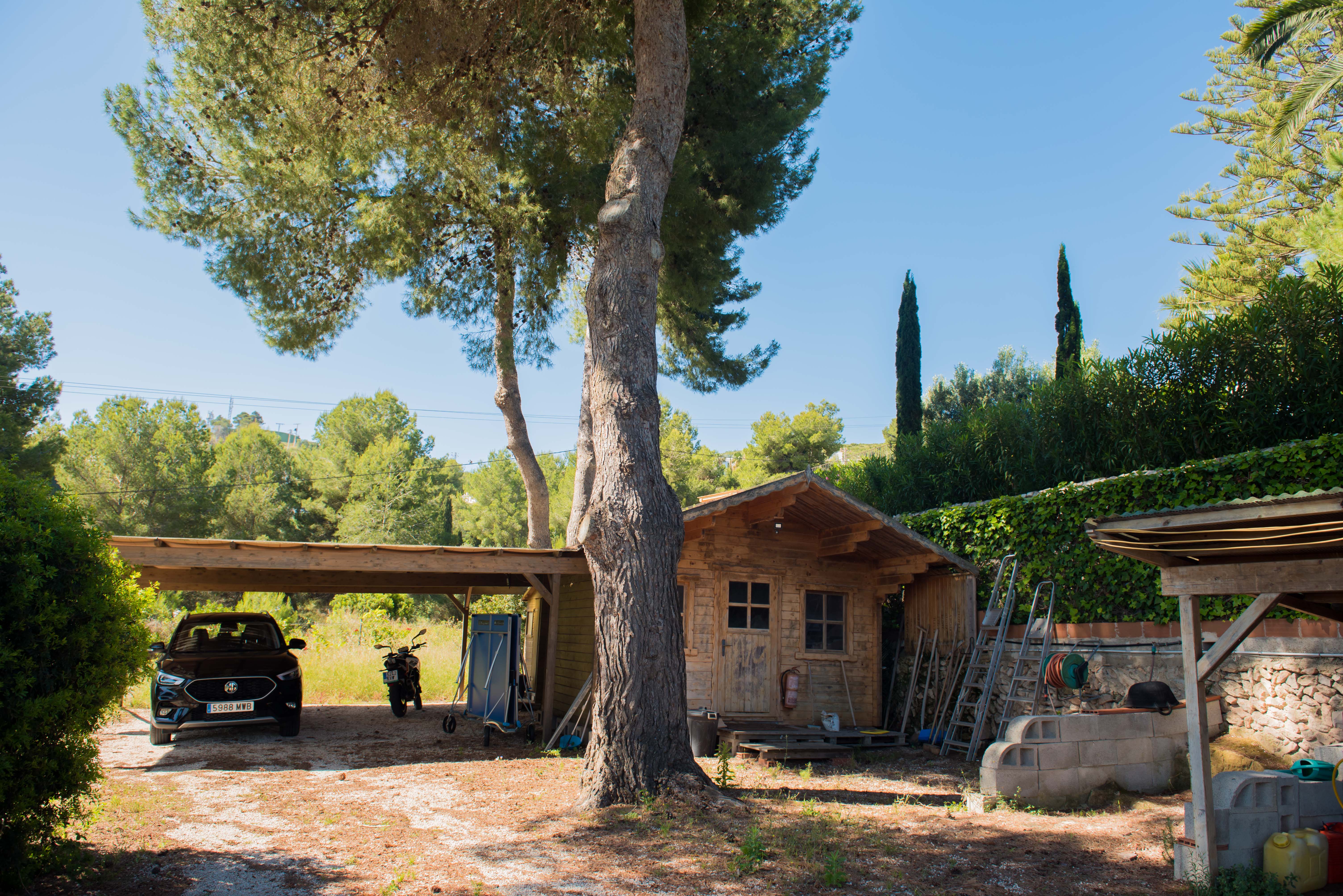 Blick auf den Holzschuppen und die überdachte Garage, umgeben von mediterraner Vegetation und klarem Himmel.