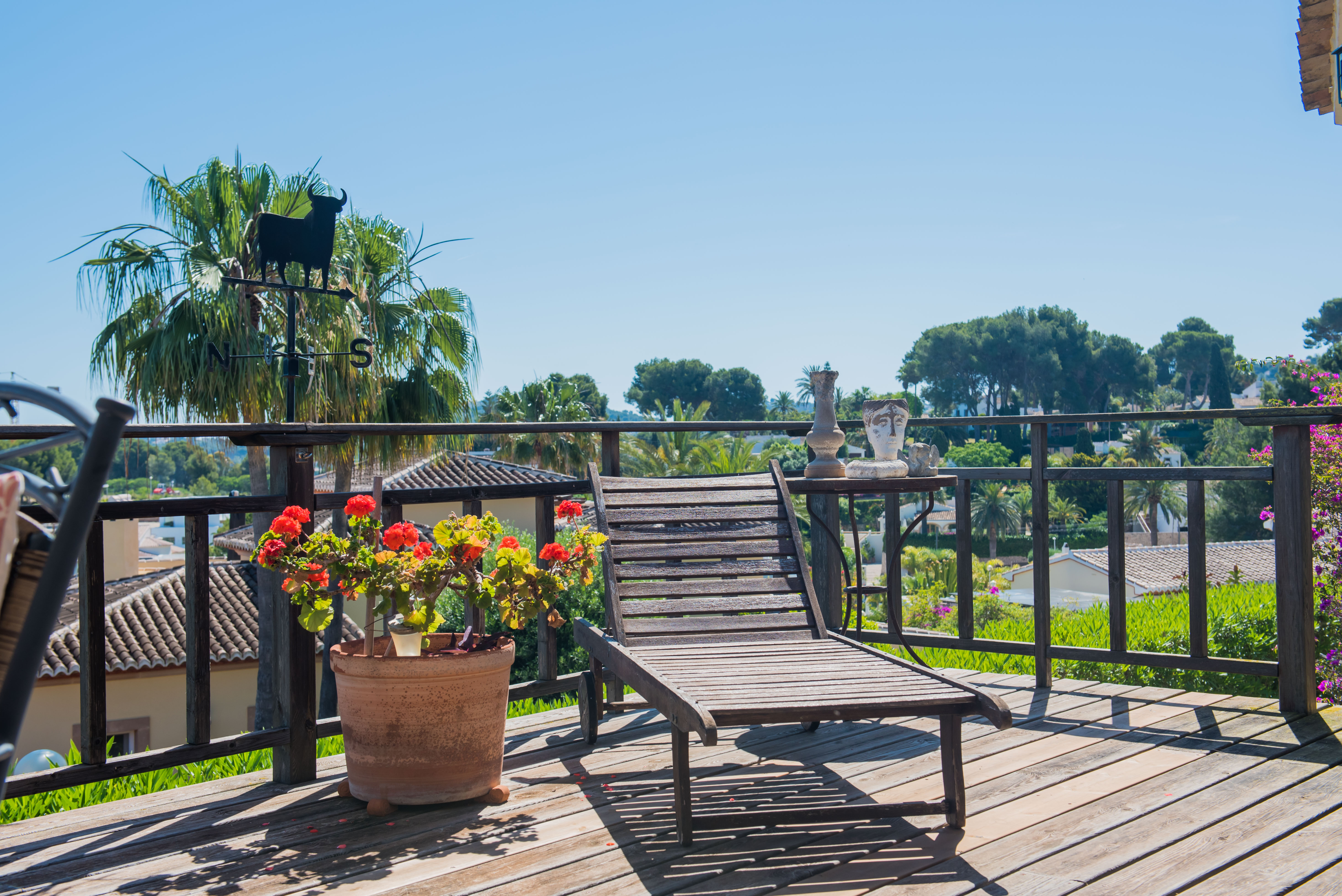 Holzterrasse mit Liegestuhl, Gartenblick und dekorativen Details wie einer Stier-Wetterfahne und Geranientöpfen.